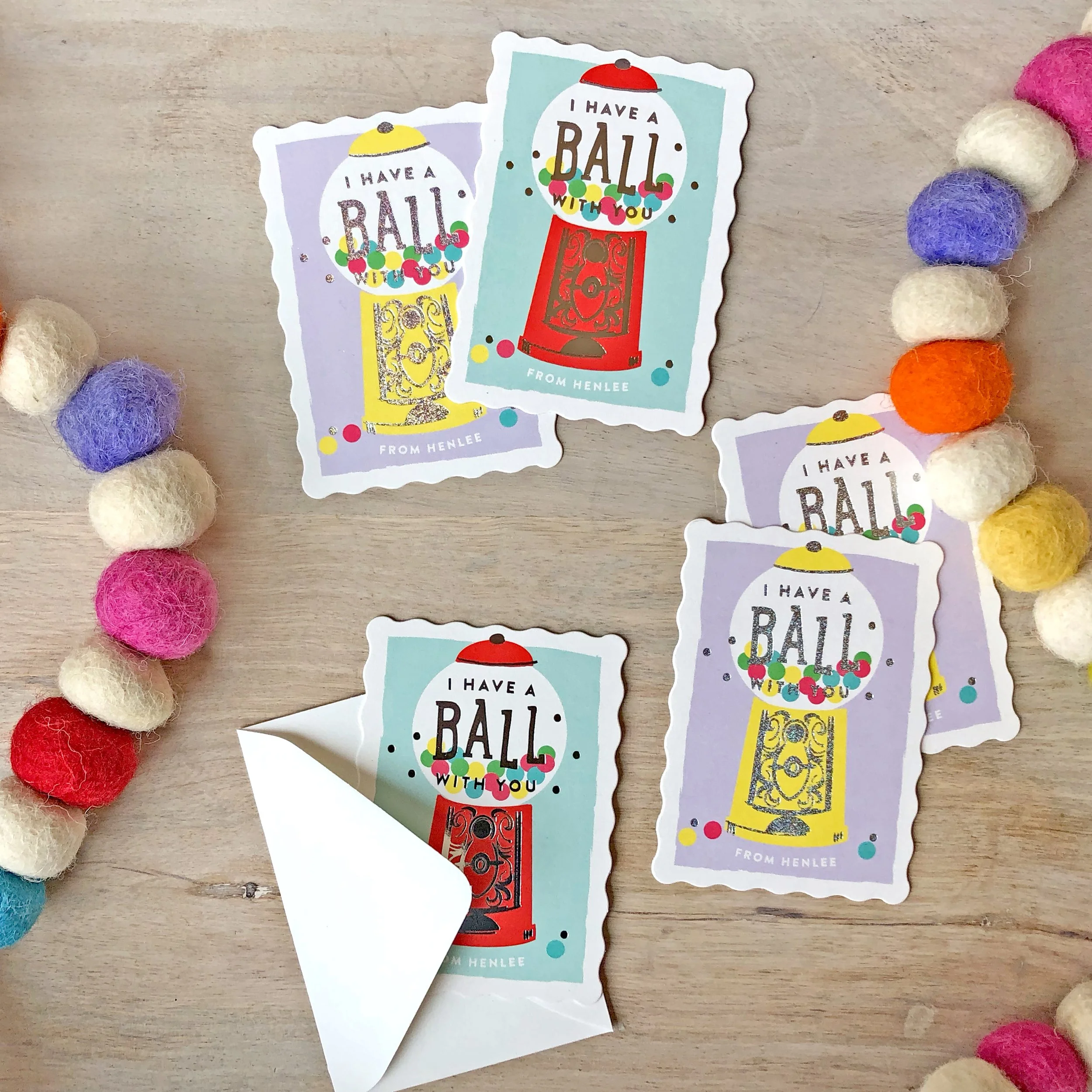 Four colorful stickers with a gumball machine design and the words "I have a ball with you" on a lavender, blue, and teal background. The stickers are scattered on a light wooden surface surrounded by a multicolored felt ball garland.