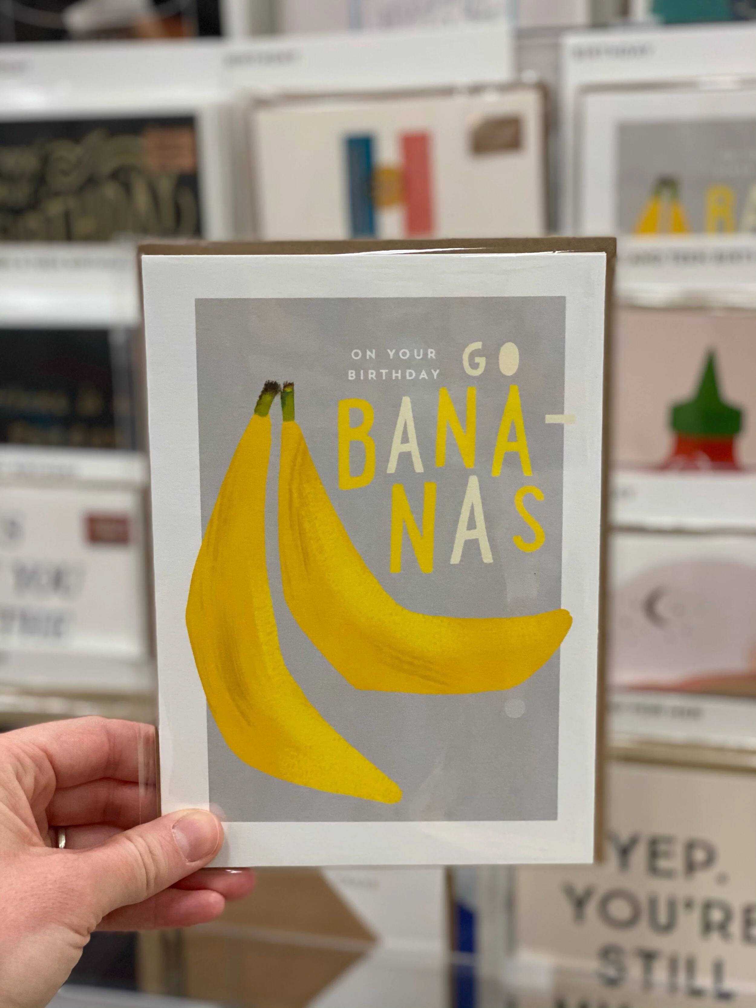 A birthday greeting card with yellow bananas and the text 'On your birthday go bananas' on a gray background, being held in a person's hand.