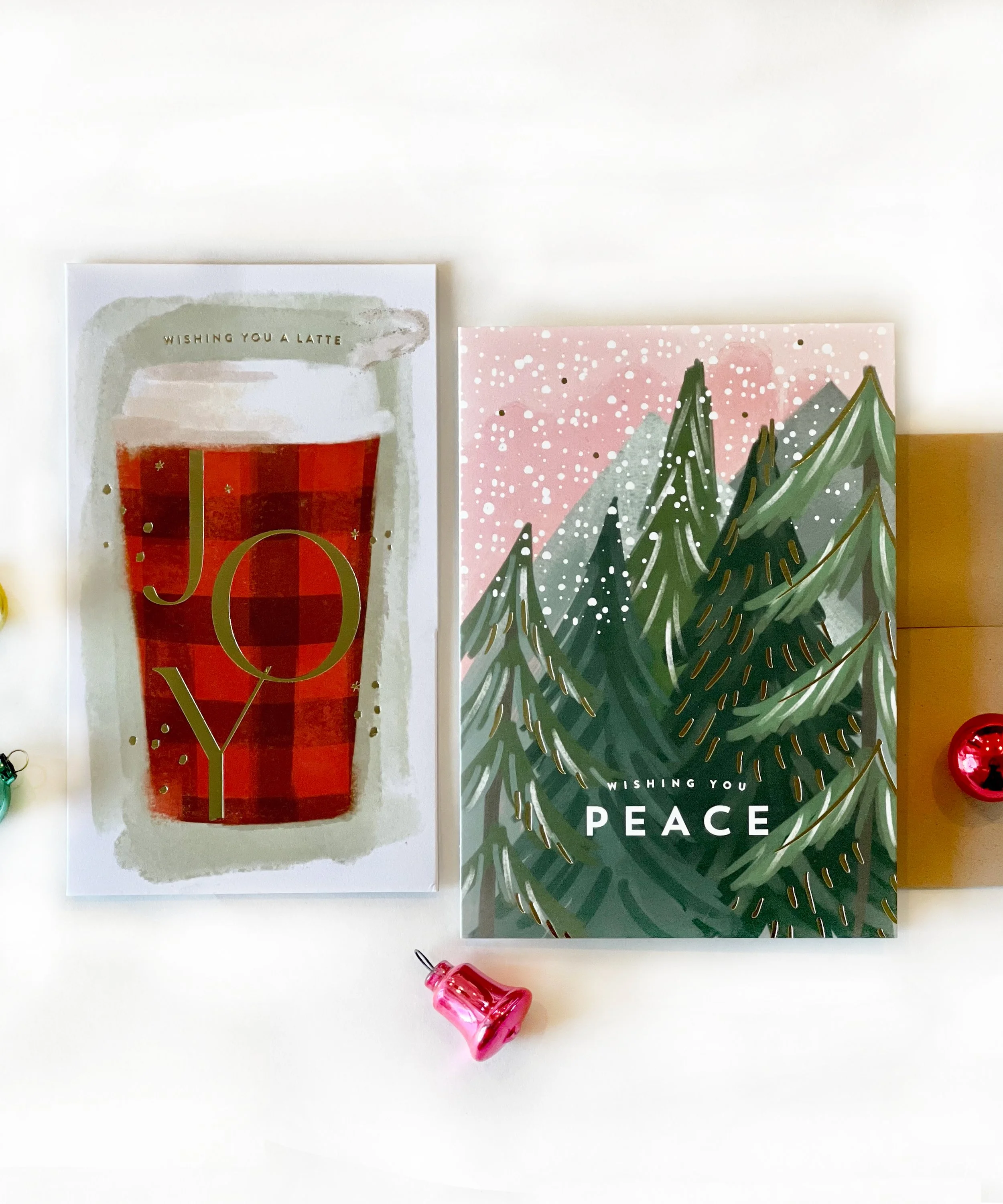 Two holiday greeting cards, one with a red latte drink and the words 'Wishing you a latte joy,' and the other with Christmas trees and the words 'Wishing you peace,' surrounded by small ornaments.