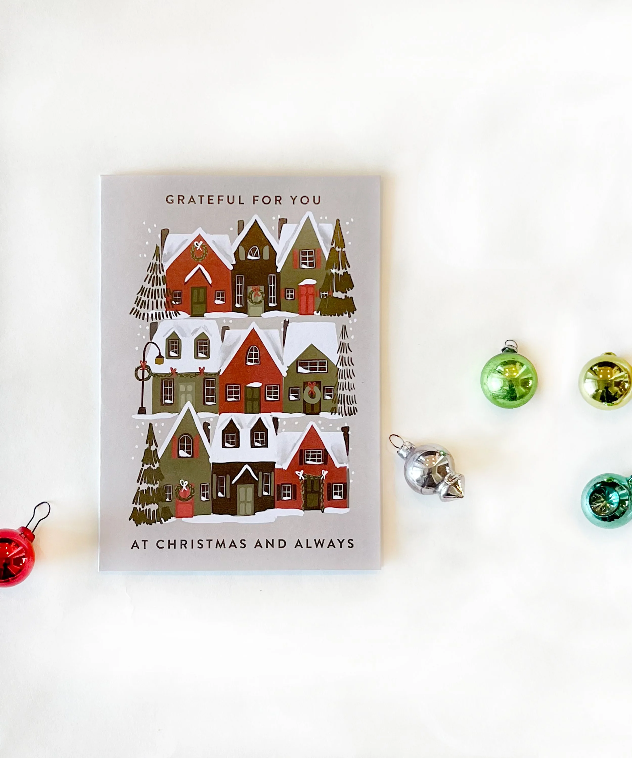 A Christmas card with illustrations of houses and trees, with the message "Grateful for you at Christmas and always," surrounded by four shiny Christmas ornaments in green, silver, turquoise, and red on a white background.