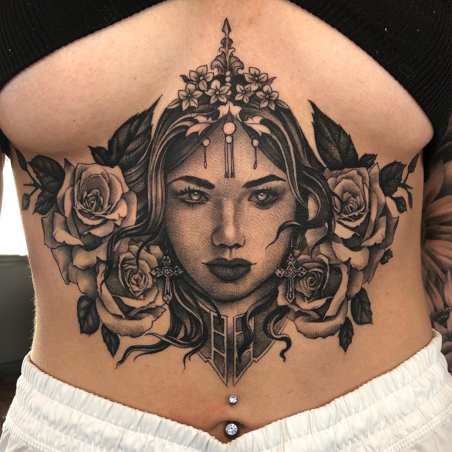 Tattoo of a woman's face with roses and floral elements on her hair, wearing earrings and a decorative collar, on a person's abdomen.