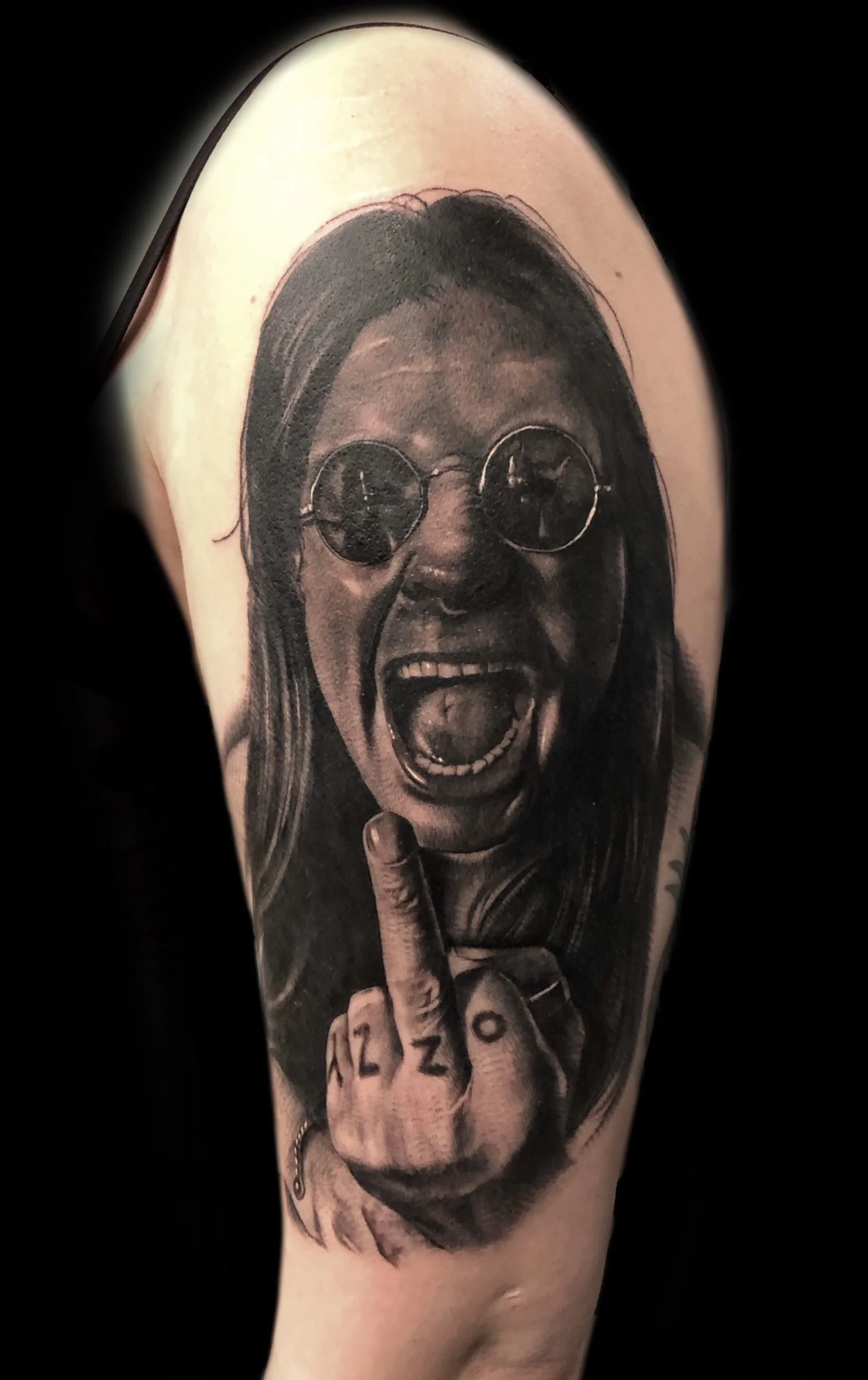 Tattoo of a woman with long hair, wearing sunglasses, shouting with her mouth open, and showing her middle finger, with the numbers '1 2 0' tattooed on the finger.
