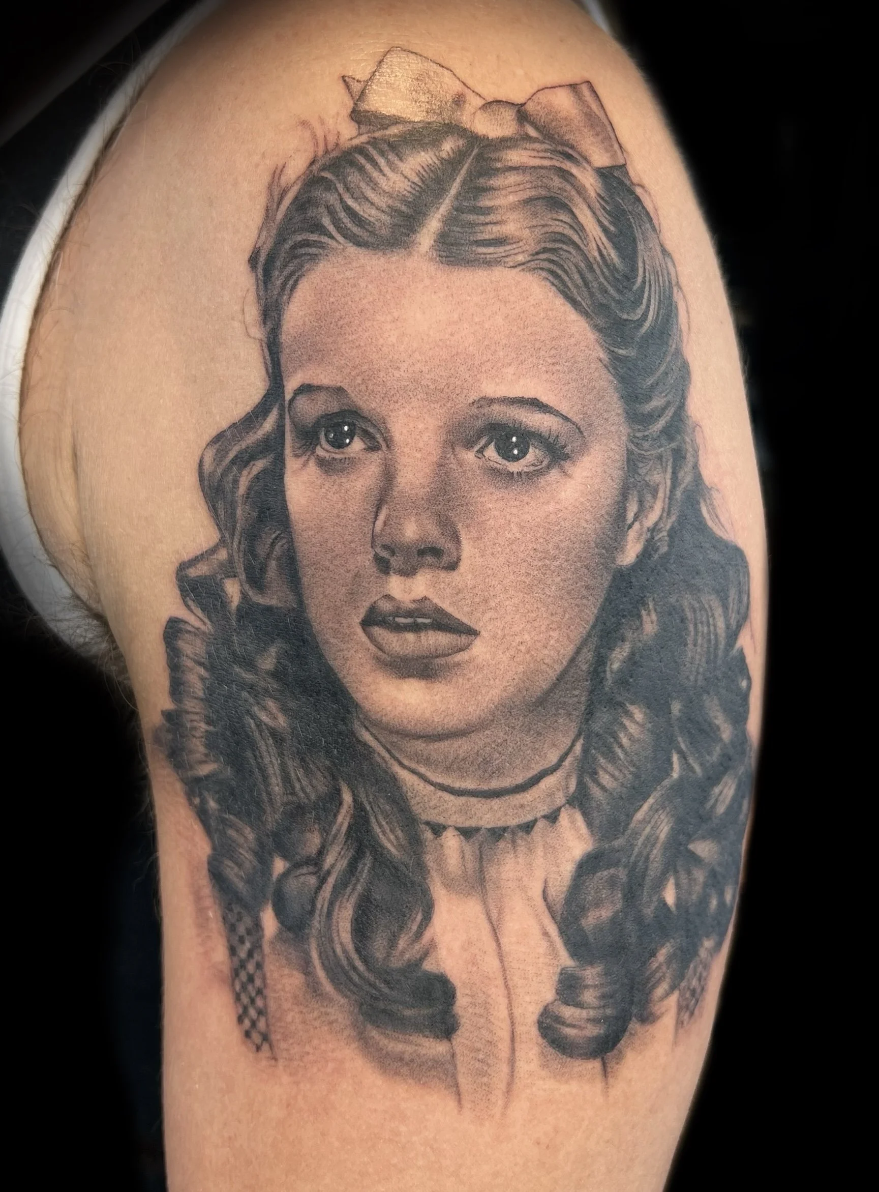 Black and gray tattoo of a realistic woman's face with wavy hair, a bow, and a choker necklace on upper arm.