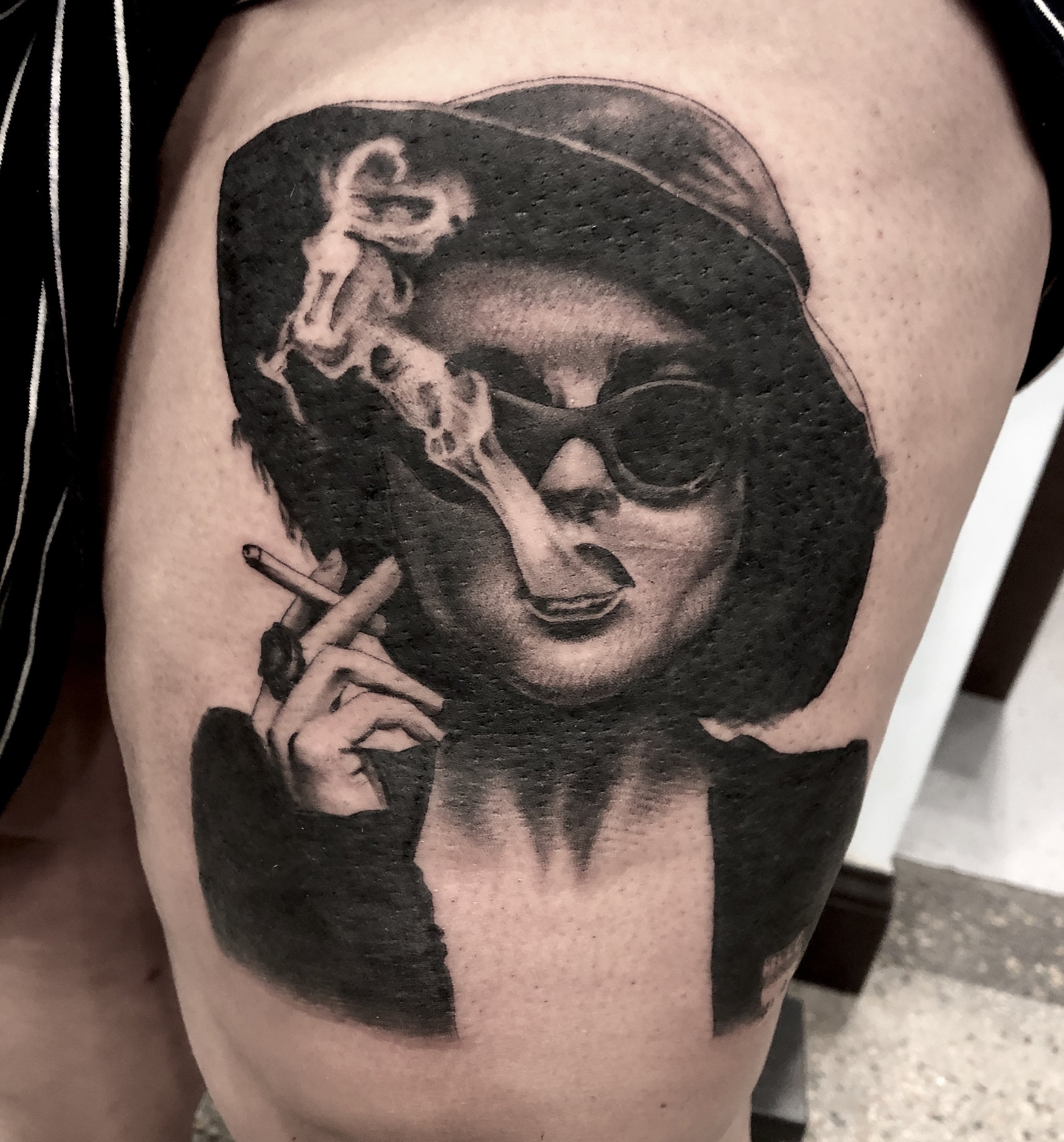 Black and gray tattoo of a woman with short hair wearing sunglasses, smoking a cigarette, and holding a smoking gun.