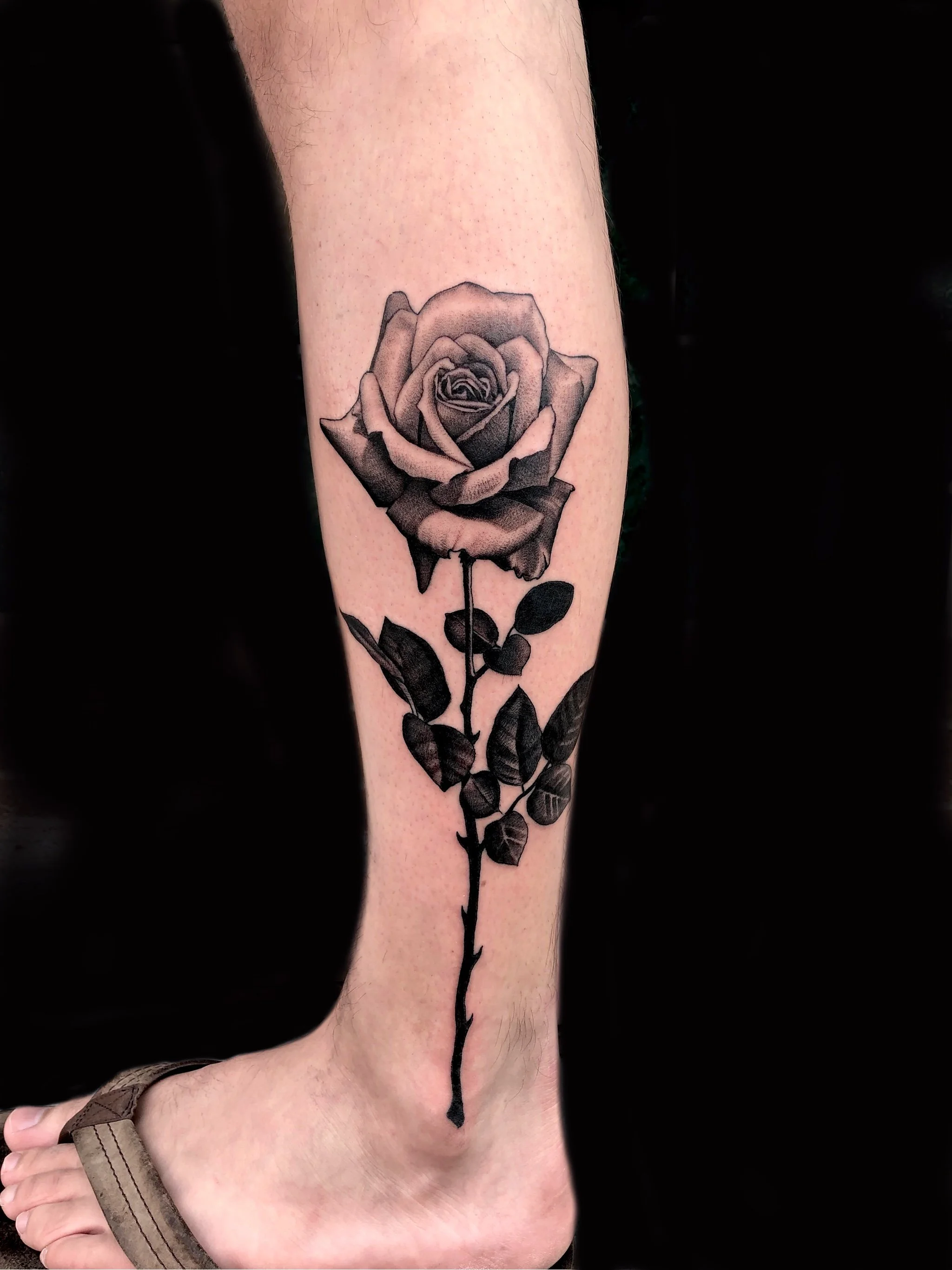 A black and gray tattoo of a detailed rose flower with leaves on a person's lower leg.