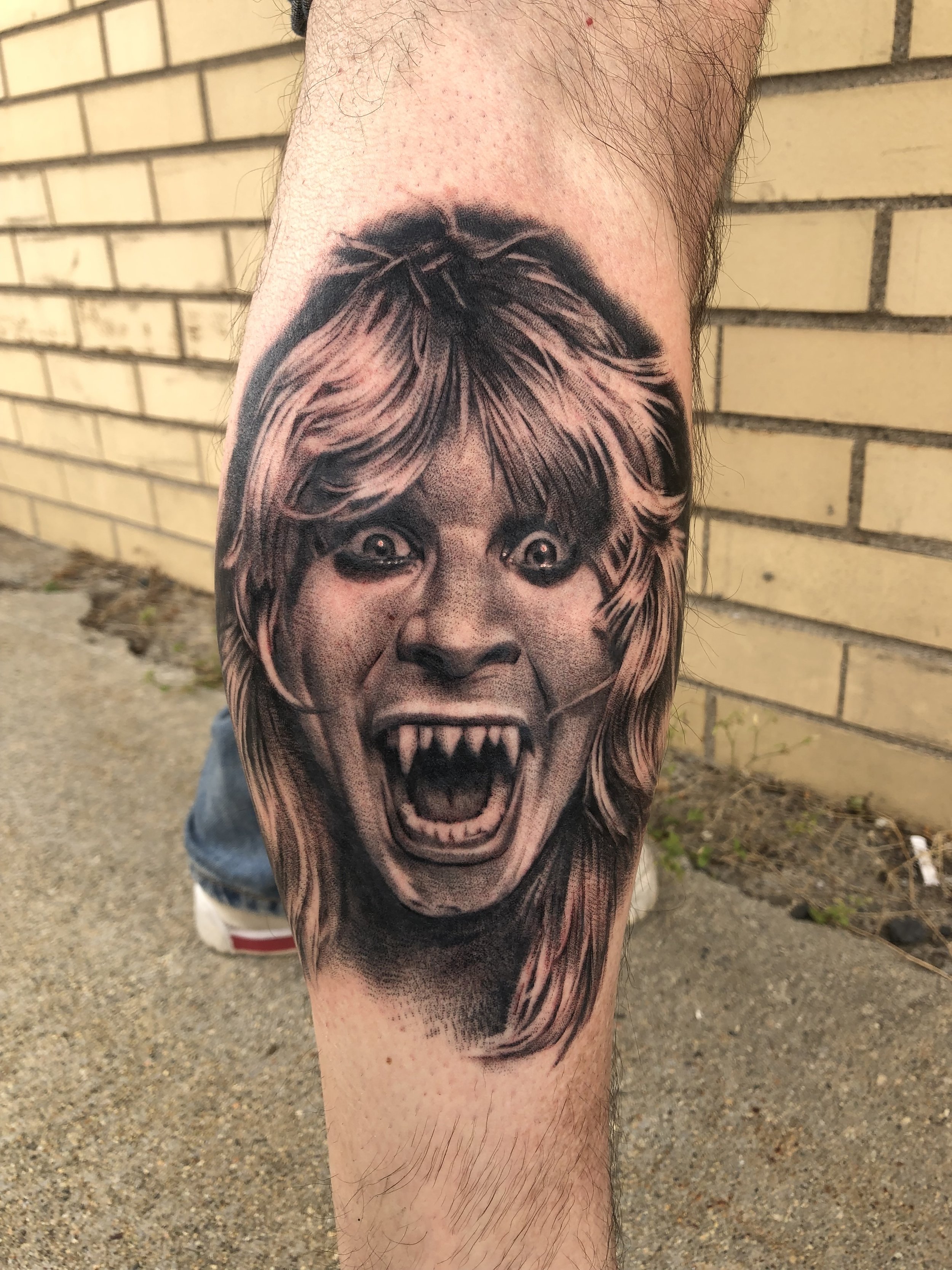 A tattoo of a frightening, monstrous face with sharp teeth and wide-open mouth on a person's calf, with a brick wall in the background.
