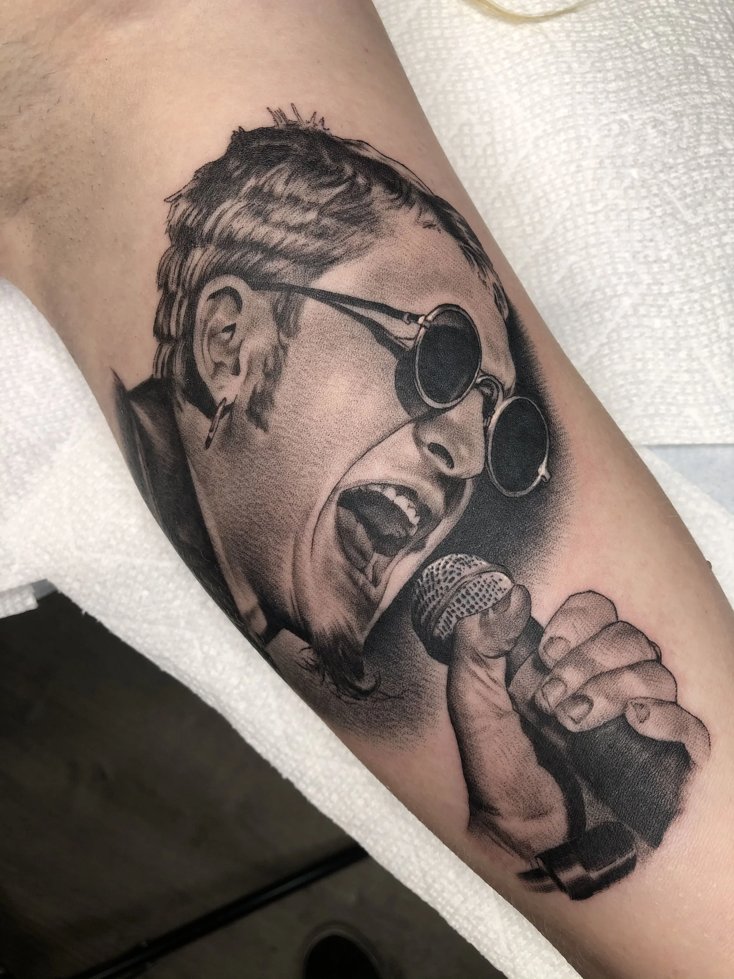A black and gray tattoo of a woman singing into a microphone, wearing sunglasses and earrings, with her mouth open and eyes closed.