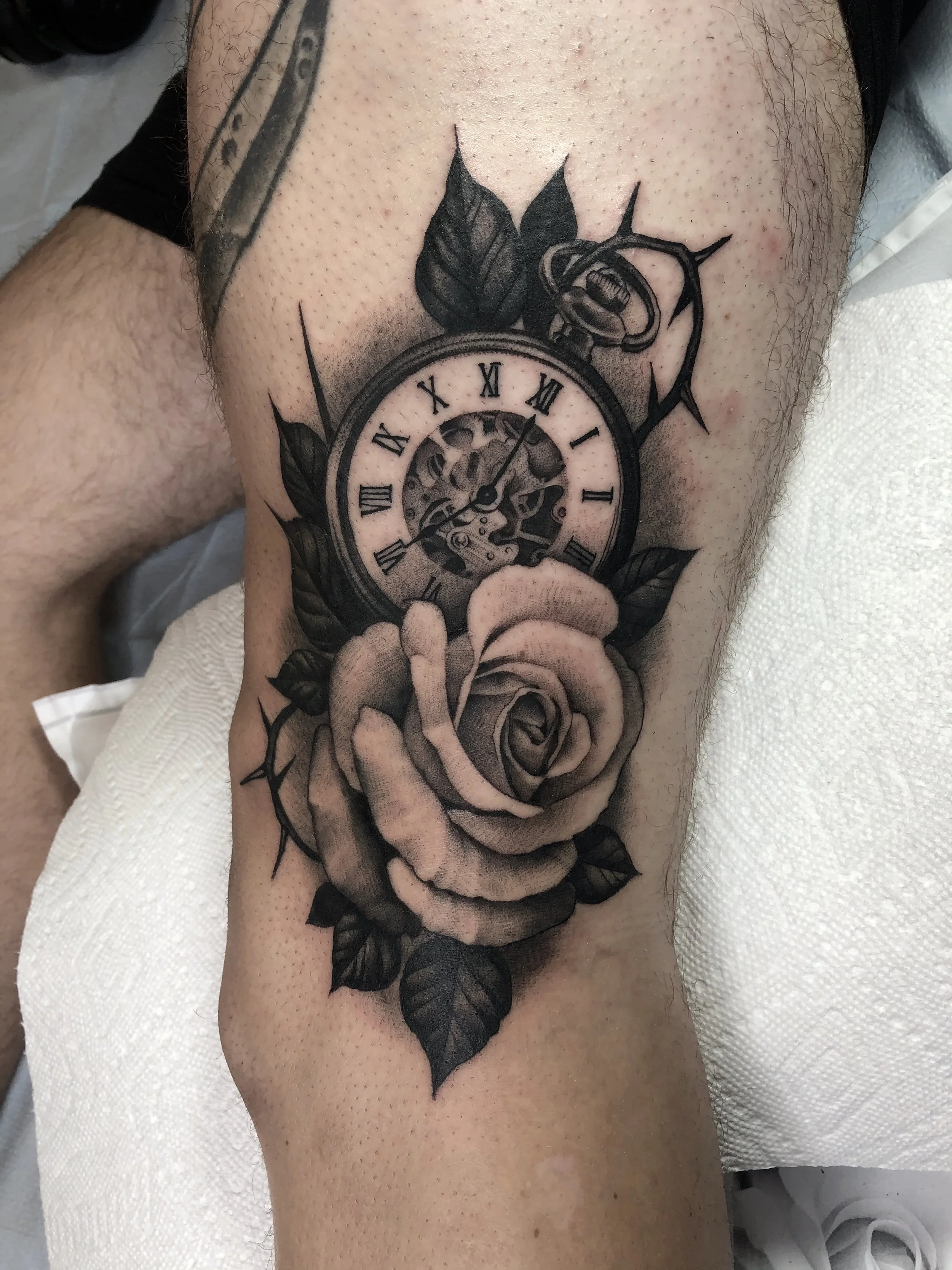 Black and gray tattoo of a pocket watch with Roman numerals, surrounded by leaves, and a large rose at the bottom, on a person's thigh.