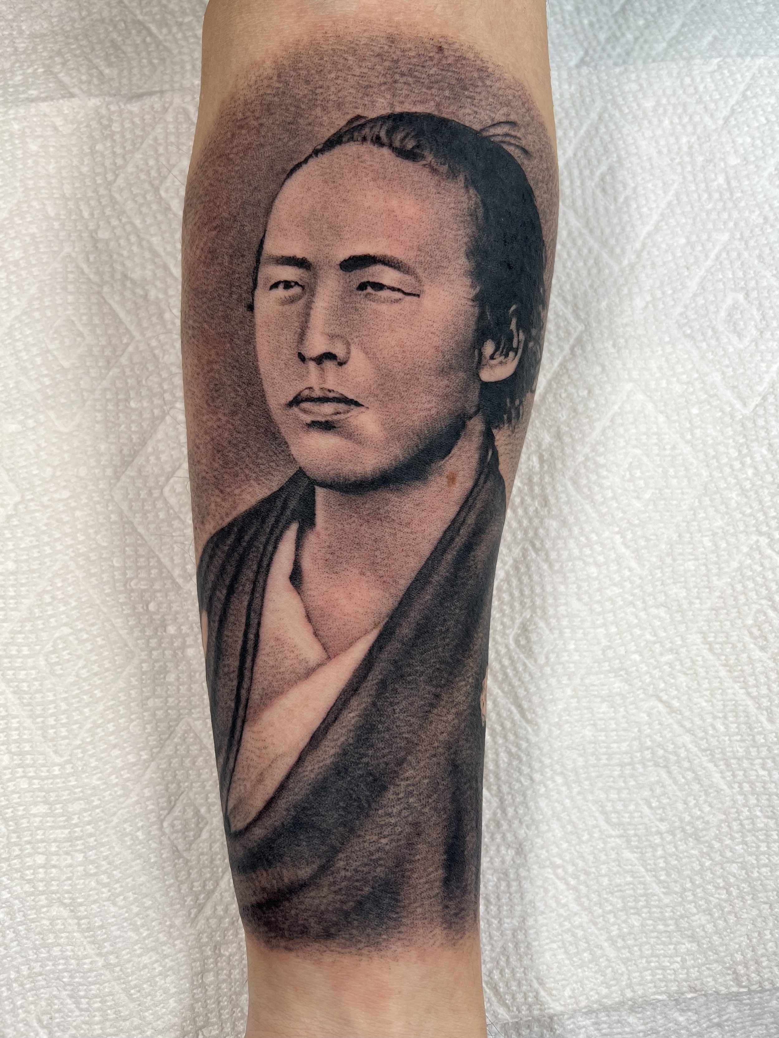 Realistic black and gray tattoo of a man with slicked-back hair, wearing traditional clothing, on a person's forearm.