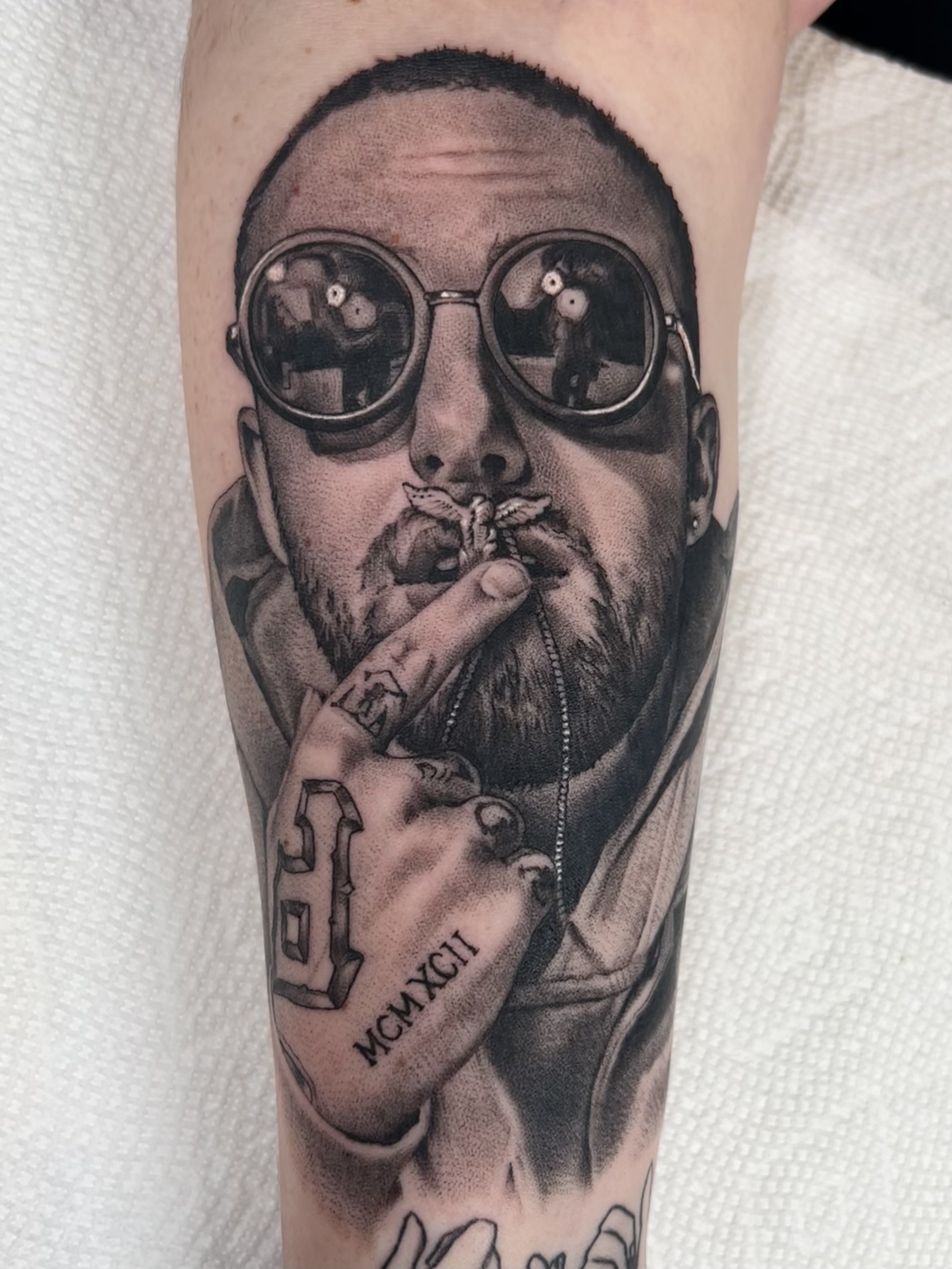 Black and gray tattoo of a man wearing sunglasses, holding a finger to his lips with a tattooed hand that shows a digital clock reading 10:10 and Roman numerals. The man has a beard and short hair.