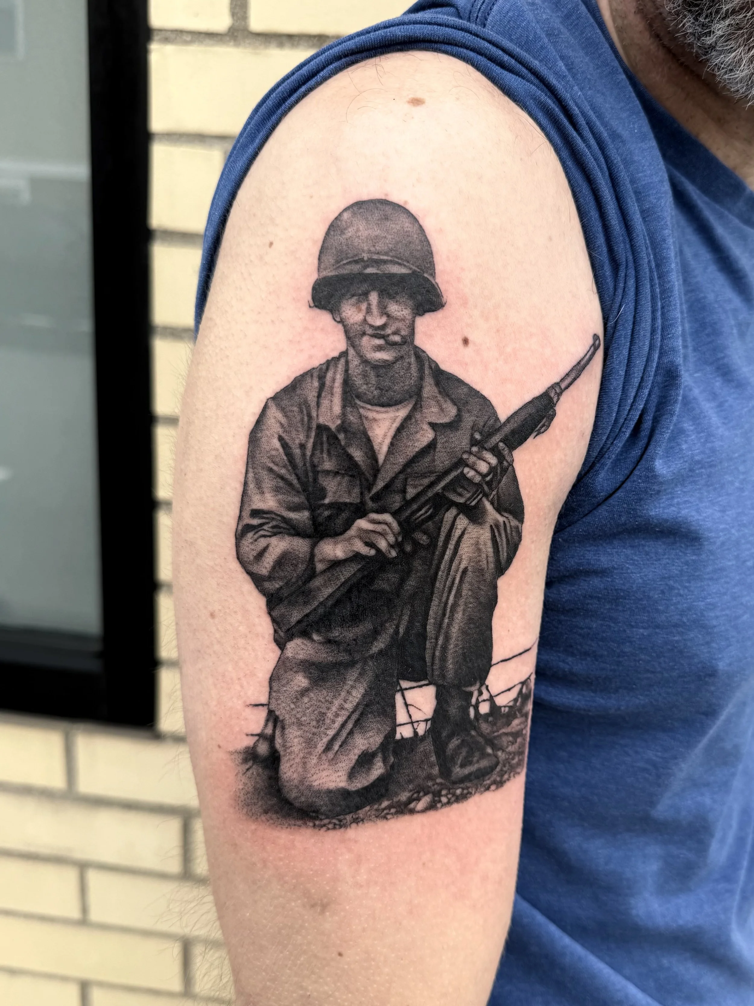 Black and gray tattoo of a kneeling soldier in military uniform holding a rifle, on a person's upper arm.
