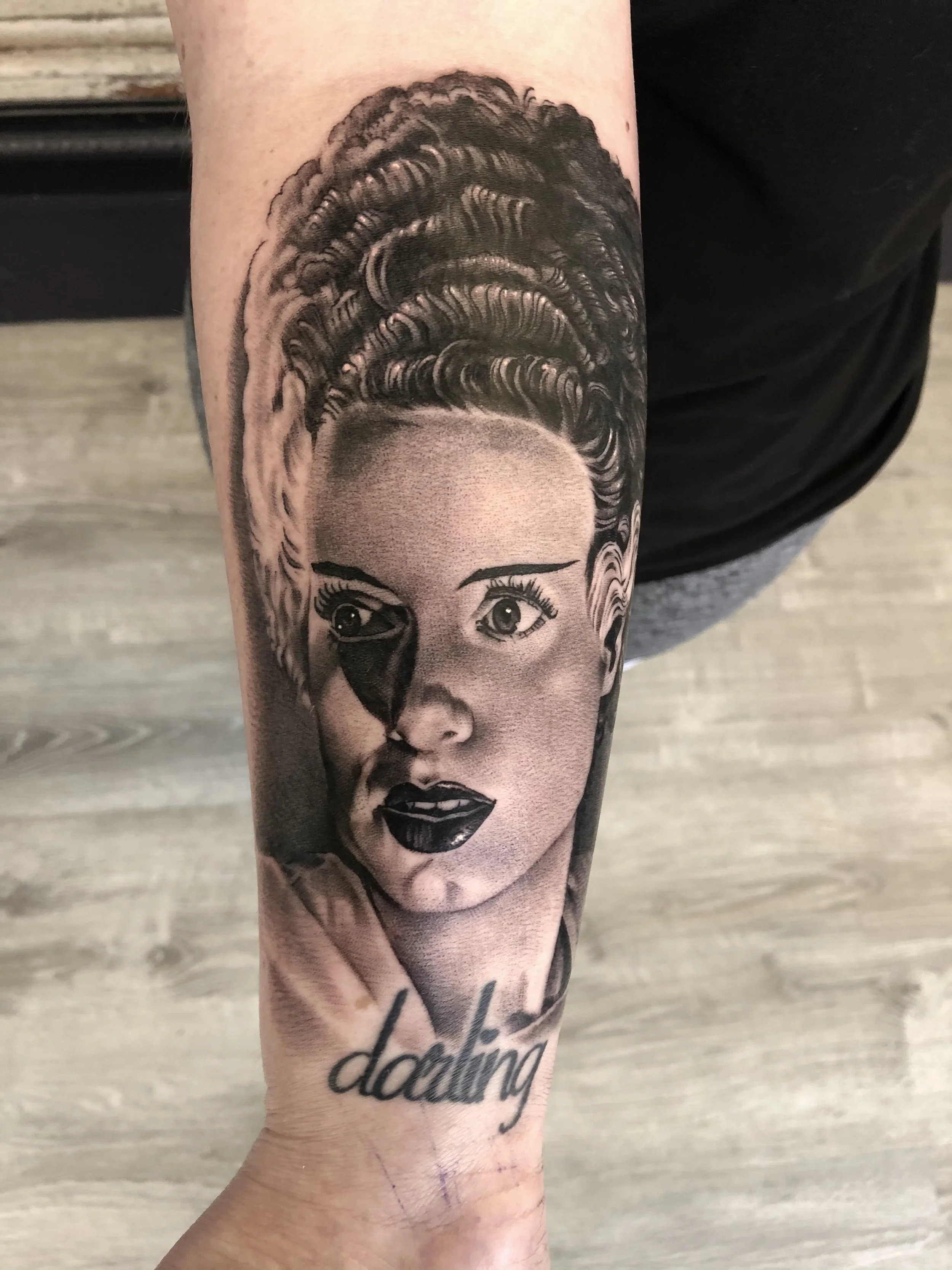 Black and gray tattoo of a woman's face with detailed hair and makeup, with a stylized black tear under her left eye, and the word 'darling' written in cursive below the portrait.