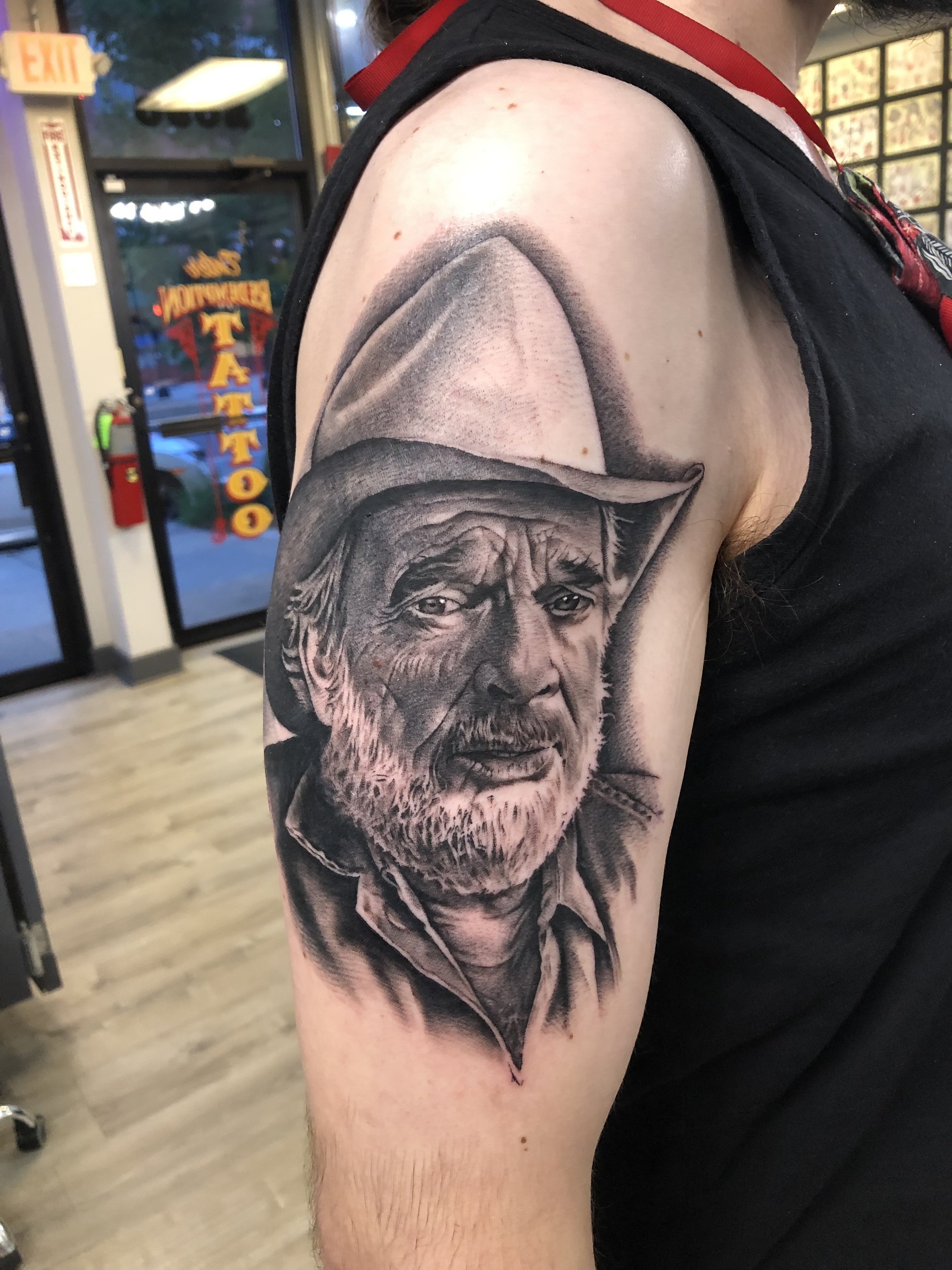 Black and gray realistic tattoo of an elderly man with a beard and mustache wearing a cap on the upper arm.