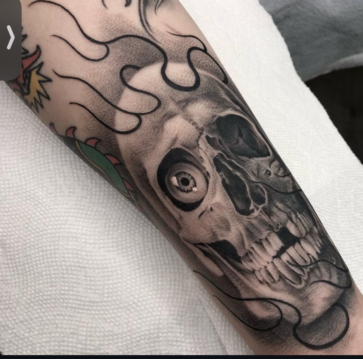 Black and gray tattoo of a skull with a detailed eye, teeth, and surrounding decorative elements on a person's arm.
