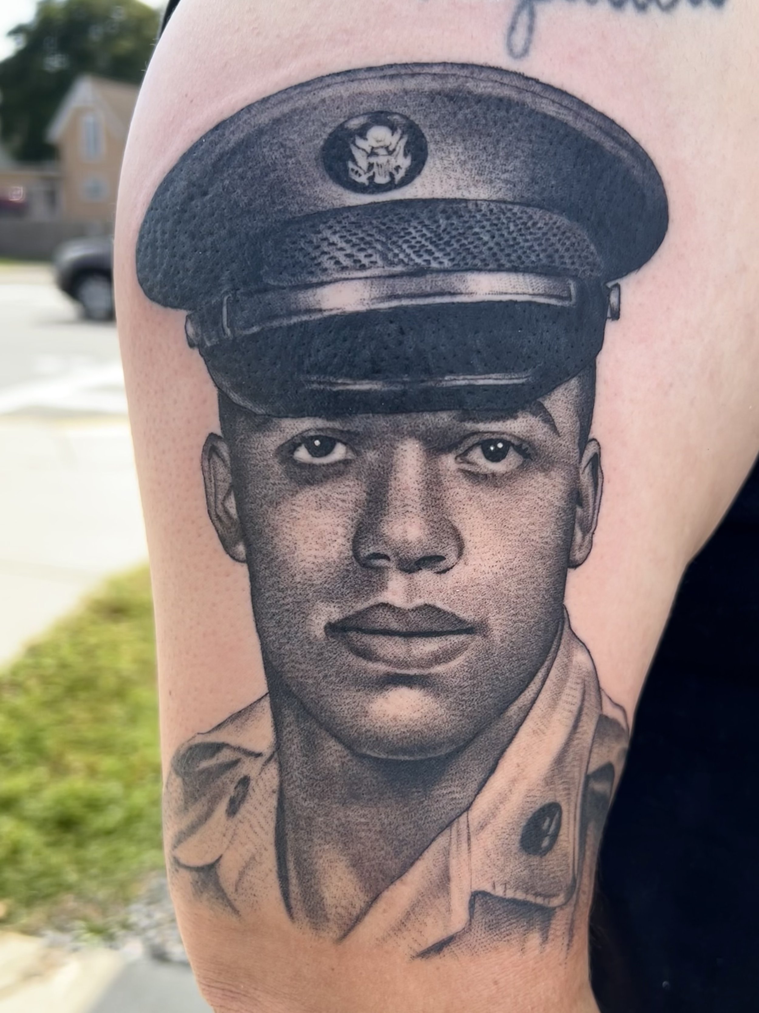 Black and gray tattoo of a young man in a military uniform wearing a military hat with an insignia, on a person's upper arm.