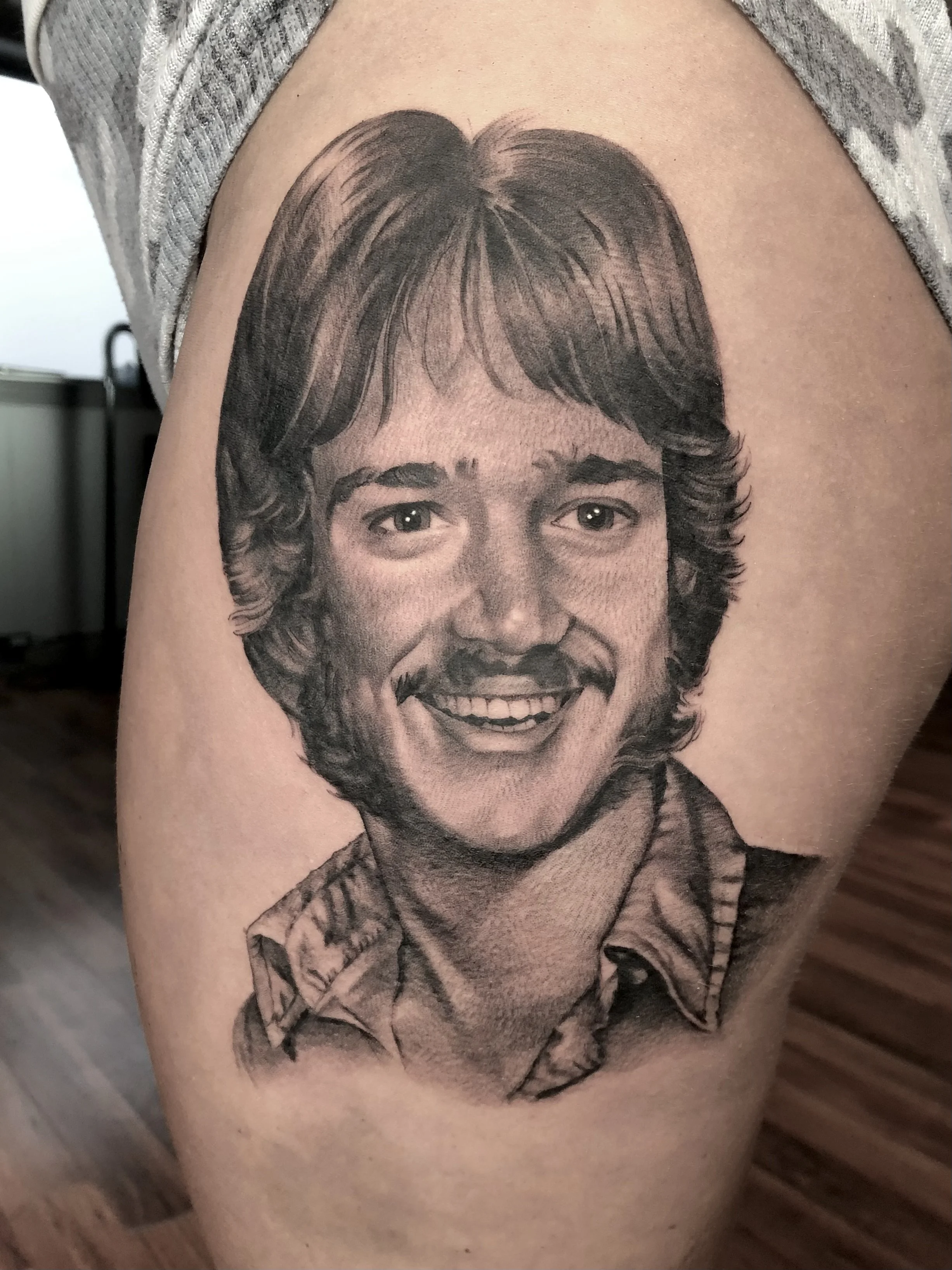 Black and gray portrait tattoo of a smiling young man with medium-length hair and a collared shirt.
