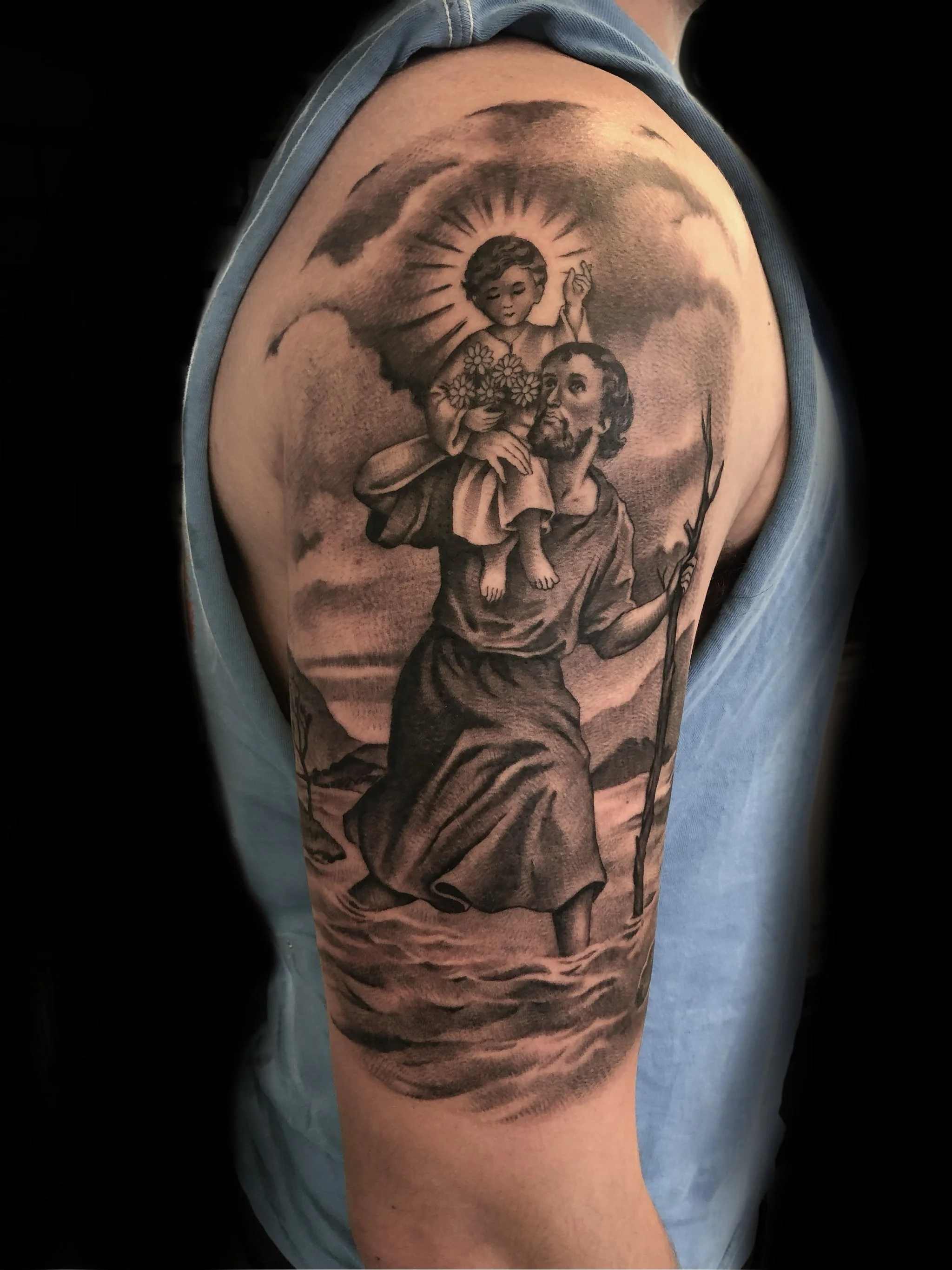 Black and gray tattoo depicting a religious scene of a man carrying a child with a halo and holding a staff, with a cloudy sky background.