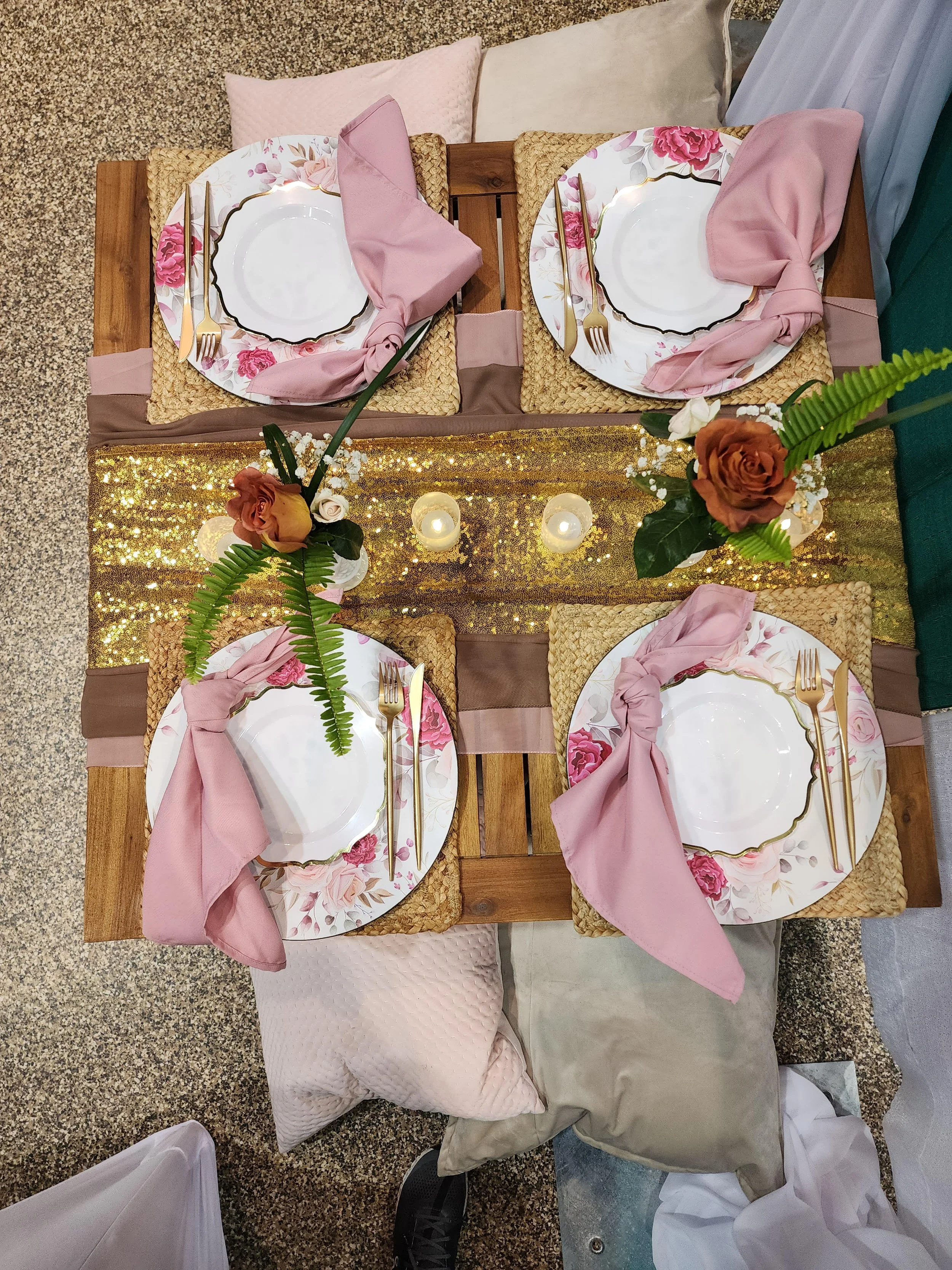 A decorated dining table set for four, with floral plates, gold utensils, pink napkins, candles, and floral centerpieces on a wood table with a gold sequin table runner.