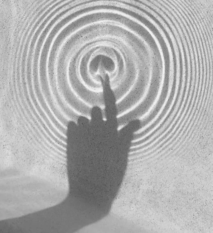 Shadow of a hand with a finger extended, touching a pointed object, creating concentric circular ripples on a surface.