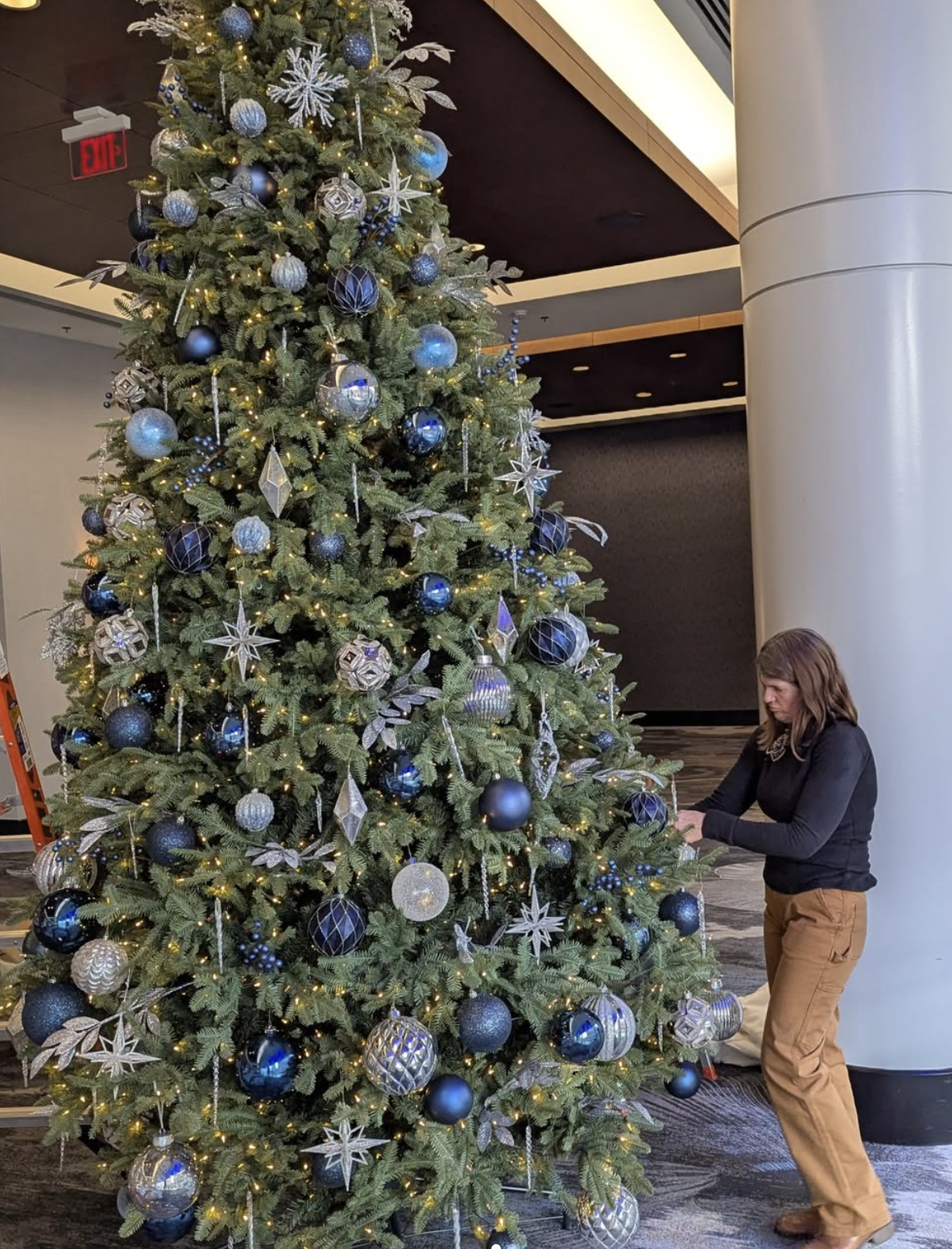Horticulturist decorating a large Christmas tree with navy and silver ornaments in a lobby.