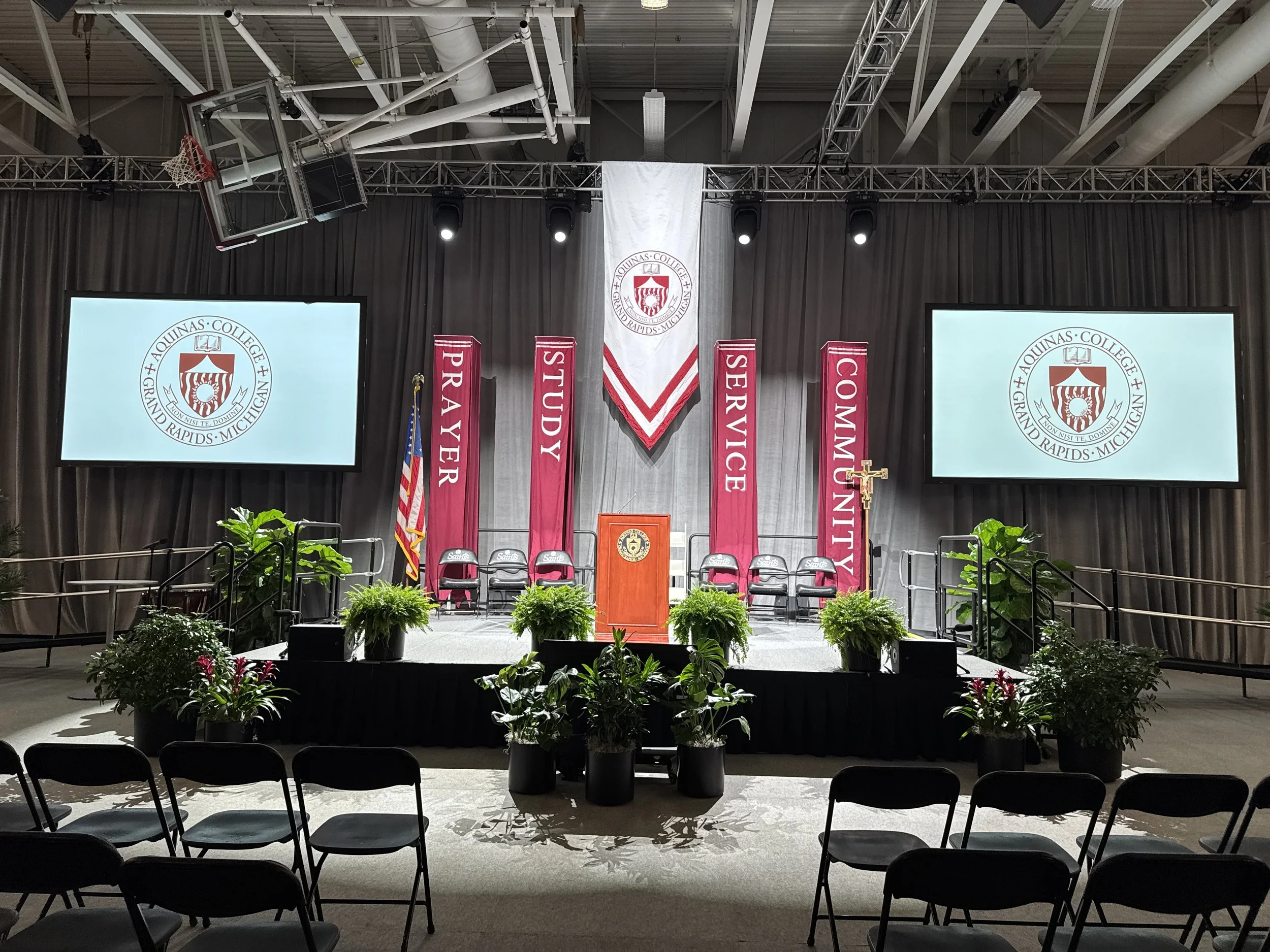 Ferns flanking a lectern on a banner-draped stage at an Aquinas College ceremony.