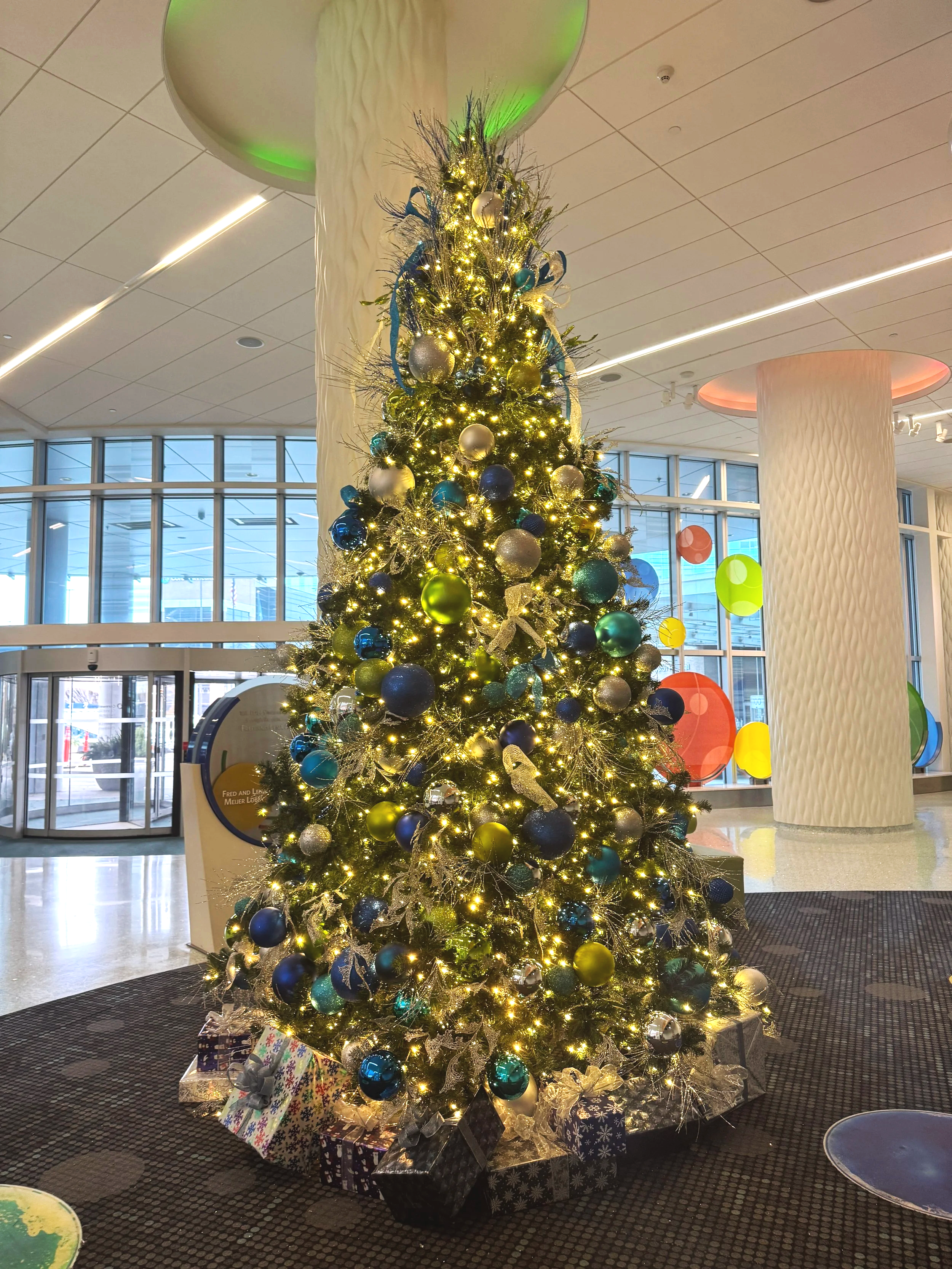 Christmas tree with blue, gold, and silver ornaments and wrapped gifts in a hospital lobby.
