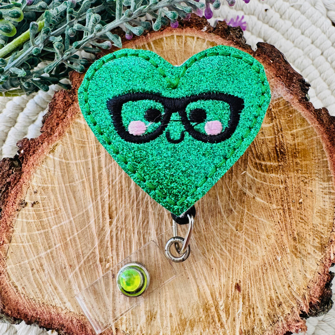 Glittery Nerdy Green Heart Badge Reel placed on a rustic wood slice with floral accents in the background.