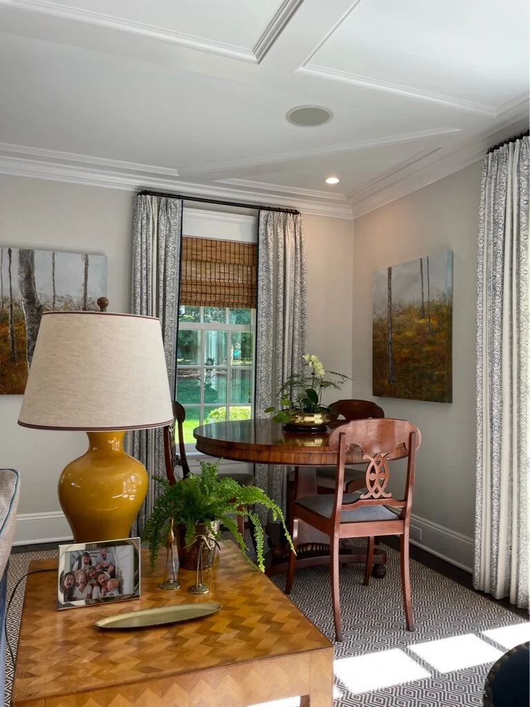 A cozy dining area with a round wooden table and three chairs, positioned near large windows with patterned curtains, artwork on the walls, a yellow table lamp, and a wooden side table with a framed family photo, a yellow vase, and green plants.