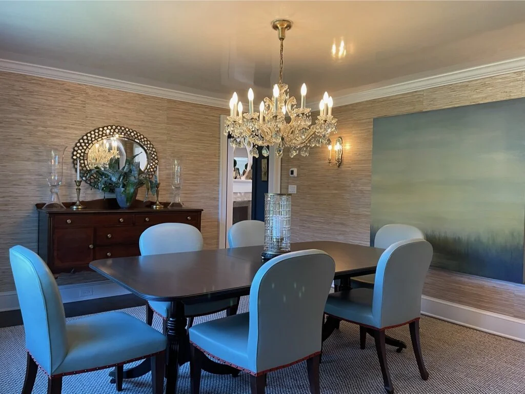 A dining room with a wooden table and six cream-colored upholstered chairs. A crystal chandelier hangs above the table, and a large cylindrical glass vase with candles is in the center. A wooden sideboard against the wall holds glass candle holders a