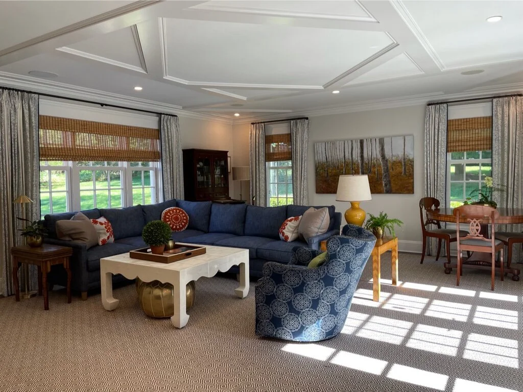 Living room with a large navy sectional sofa, patterned armchair, wooden side tables, a white coffee table, a large landscape painting, and windows with patterned curtains and bamboo blinds.