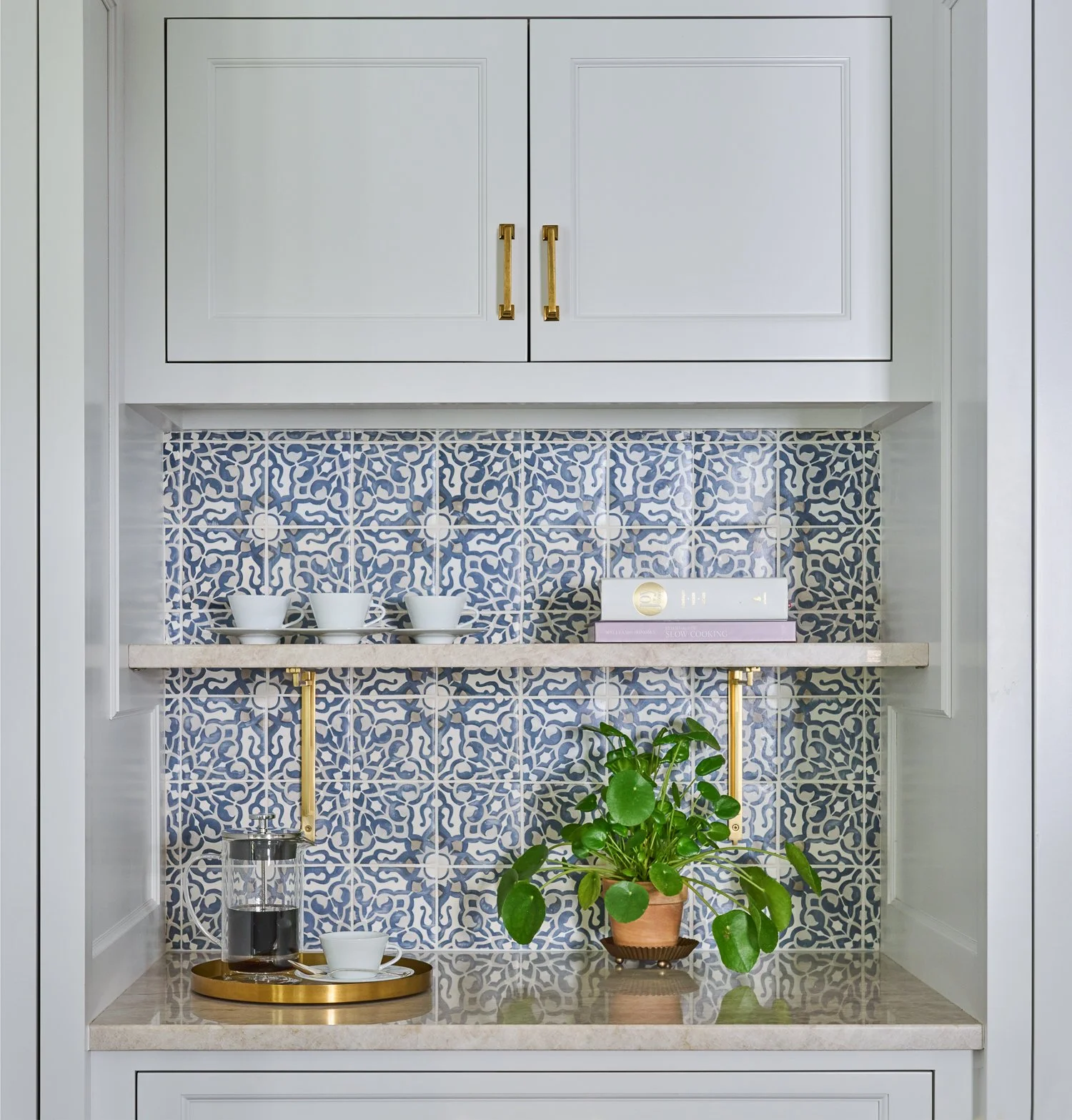 Kitchen cabinet with decorative blue and white tile backsplash, a shelf holding white cups and cookbooks, and a countertop with a potted plant and a glass coffee server with a white cup on a gold tray.