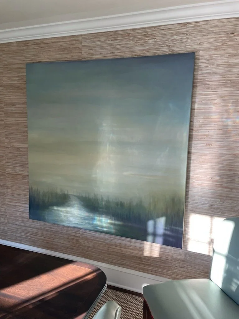 A large landscape painting of a serene natural scene with a cloudy sky, a body of water, and trees in the distance, mounted on a textured wooden wall in a room.