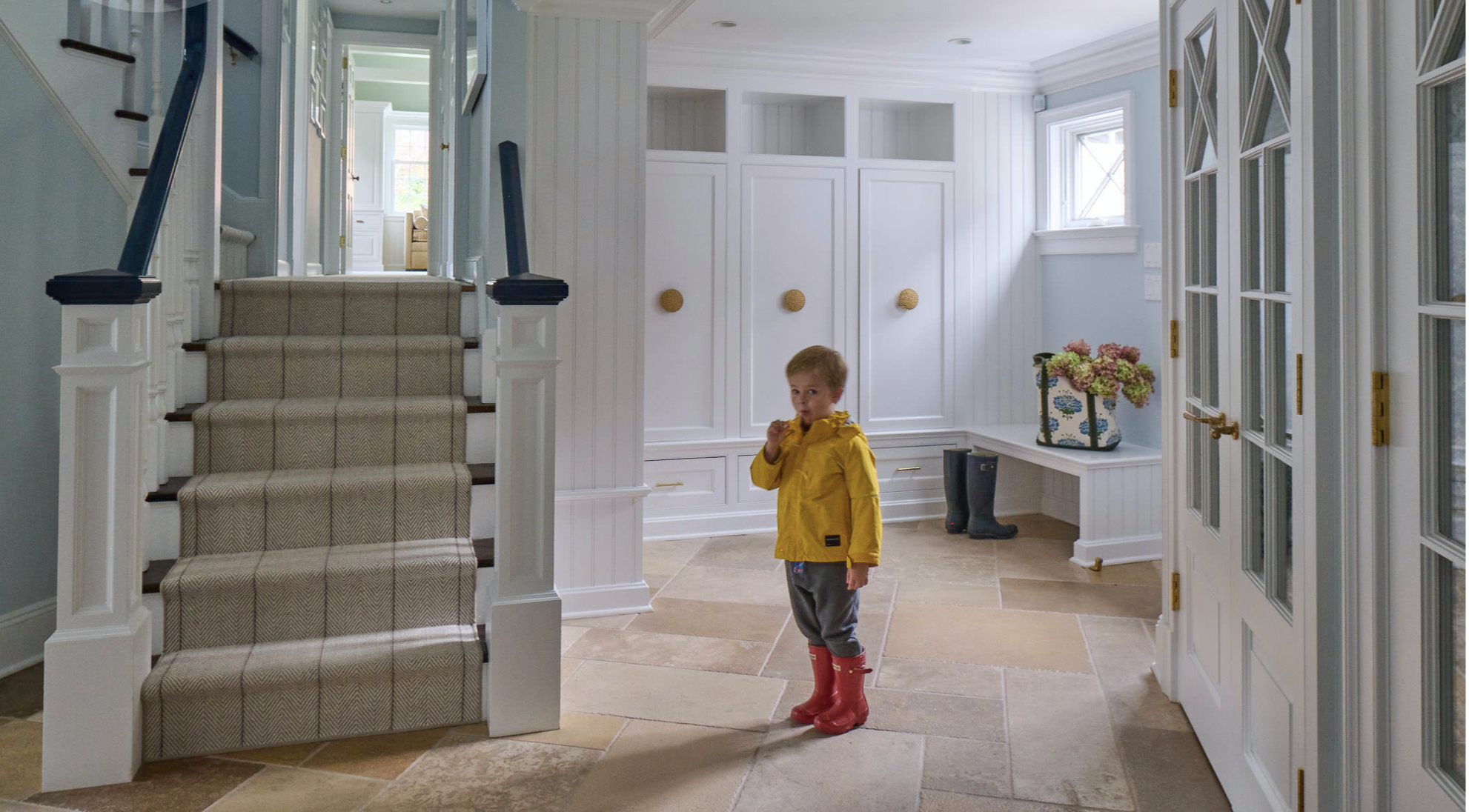 A young boy in a yellow raincoat and red rain boots stands inside a bright, spacious entryway, looking at the camera while holding a popsicle. The area features white paneled walls, built-in cabinets, and a staircase with a patterned runner rug on the stairs. There are windows allowing natural light and a bag with flowers on a bench. A pair of boots sits on the floor near the cabinet.