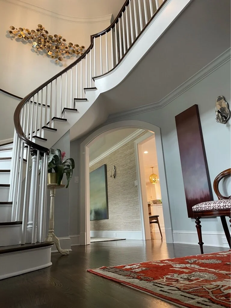 Interior view of a house entrance with a curved staircase on the left, decorated with white and dark wood balusters. A colorful abstract wall sculpture is mounted on the wall above the staircase. To the right, there is an arched doorway leading to an