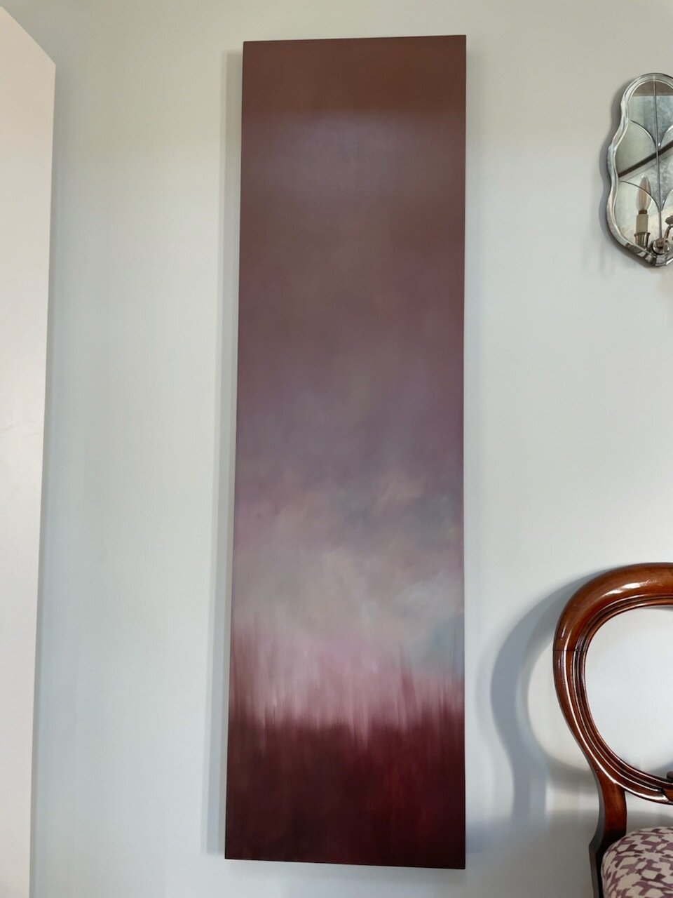 A tall, rectangular abstract painting with gradient shades of maroon, gray, and purple hanging on a white wall, with a decorative mirror and part of a wooden chair visible nearby.