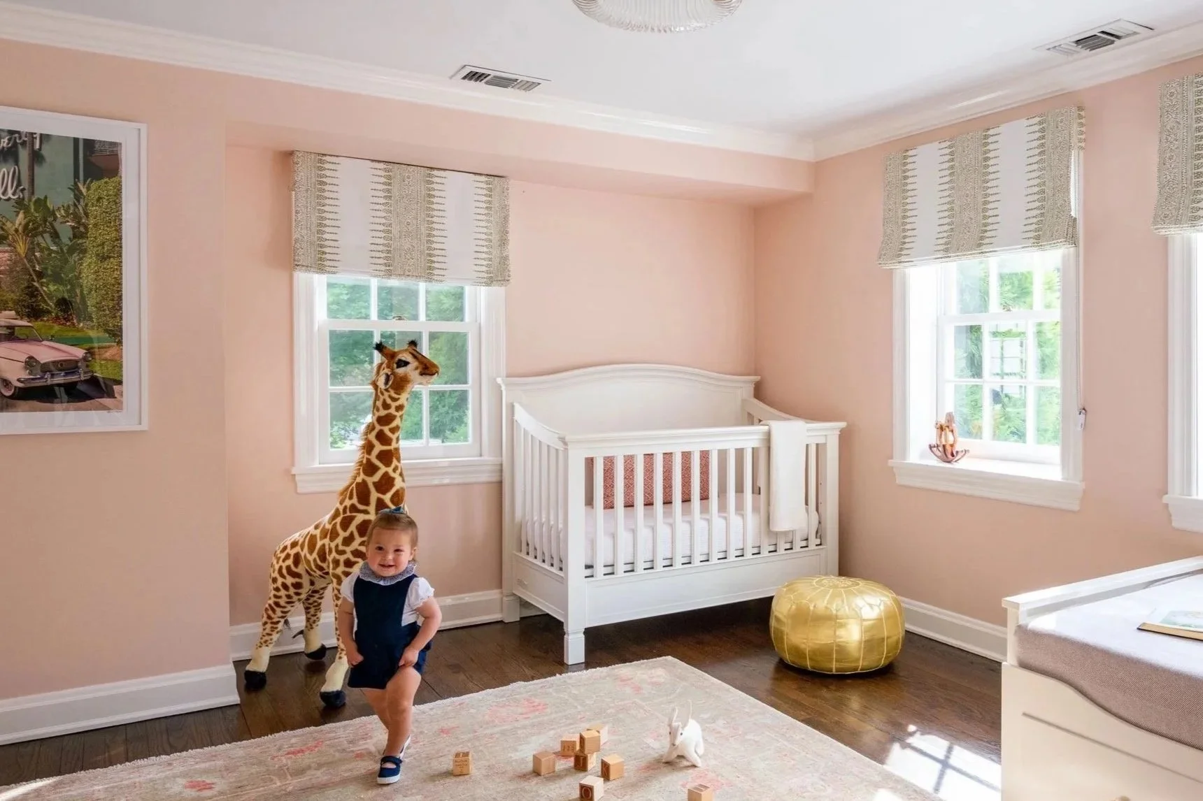 A young girl with a short-sleeve shirt and overalls playing in a nursery with a toy giraffe, wooden blocks, a bunny stuffed animal, a white crib, a yellow ottoman, and windows with patterned curtains.