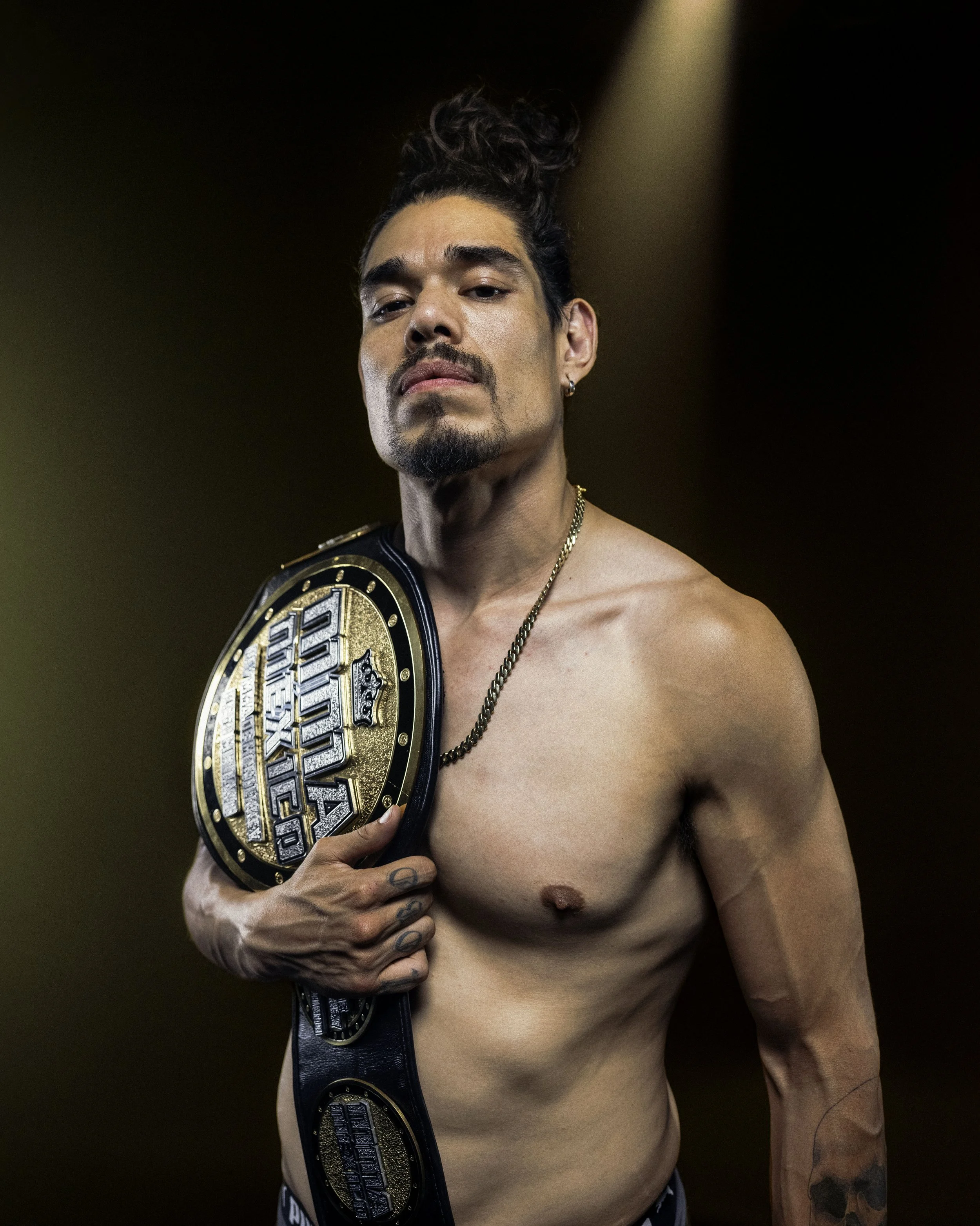 Fotografía Corporativa para Commercial y Publicidad, San Pedro Garza García, México:  Joven hombre sin camisa sosteniendo un cinturón de campeón en su hombro izquierdo, con cabello oscuro recogido y tatuajes en su brazo derecho, en un fondo oscuro.