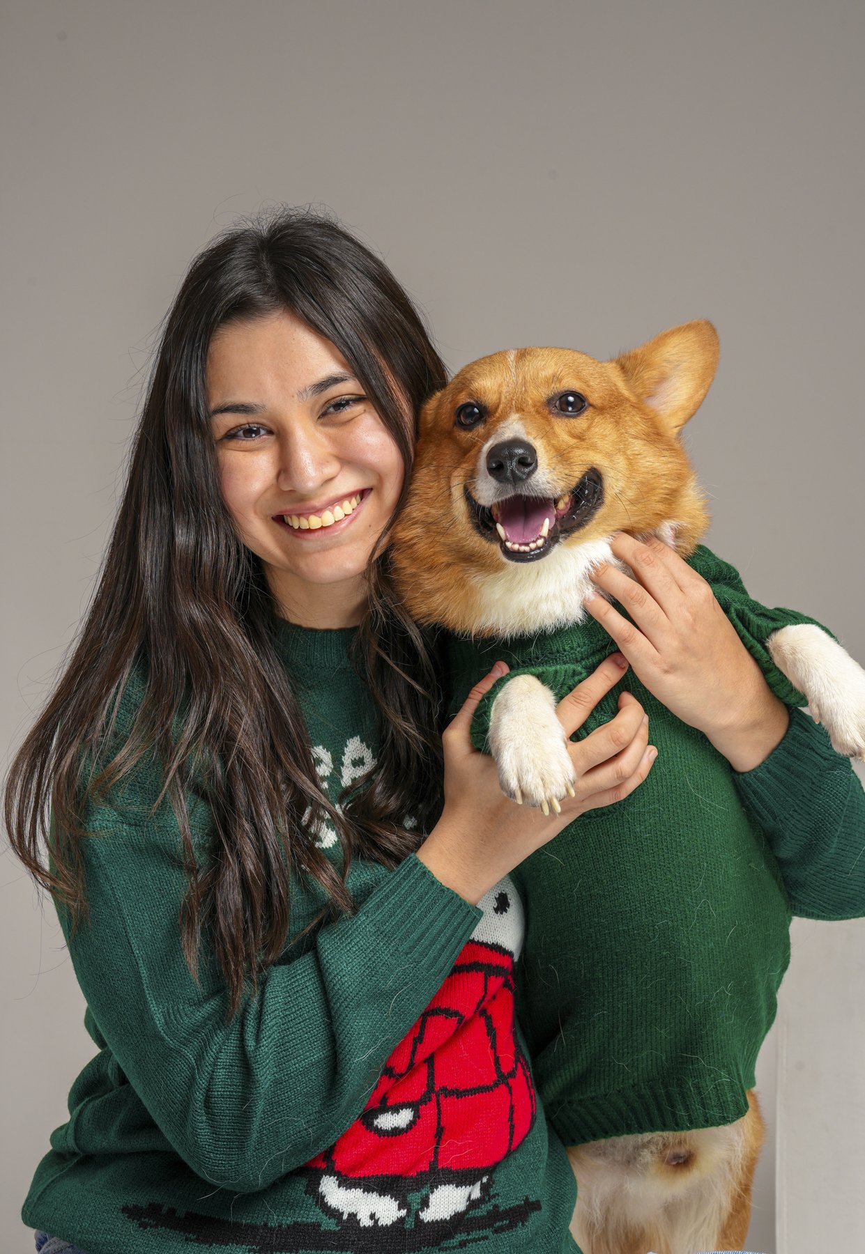 Fotografía de Mascotas: Una joven con una sonrisa brillante sostiene a un perro de raza Corgi que lleva un suéter verde. Ambos parecen felices y están frente a un fondo gris claro.
