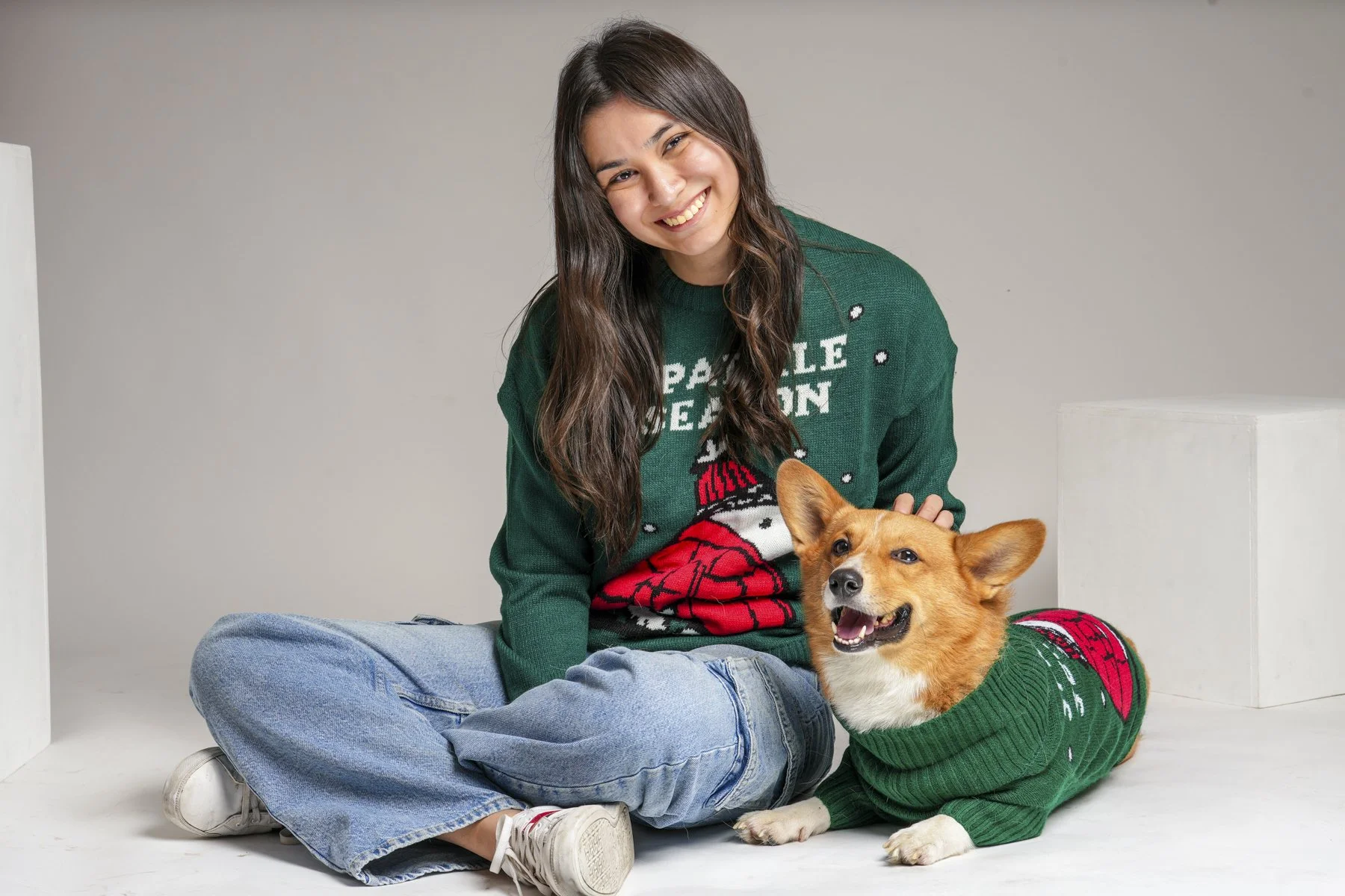 Fotografía de Mascotas: Una joven mujer con una sonrisa, vestida con un suéter verde con motivos navideños y un perro con un suéter a rayas verdes y rojos, sentados en el suelo contra un fondo gris.