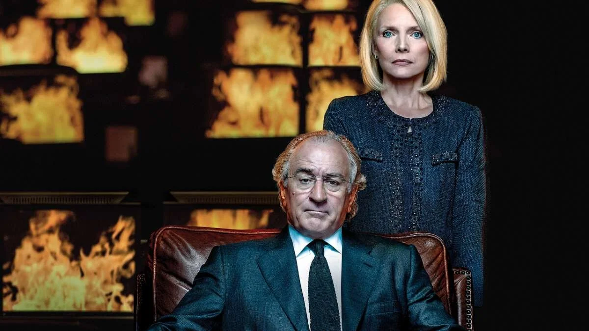 Robert de Niro in a suit and a woman in a dark blazer posing in a dimly lit room with fiery images on screens in the background.