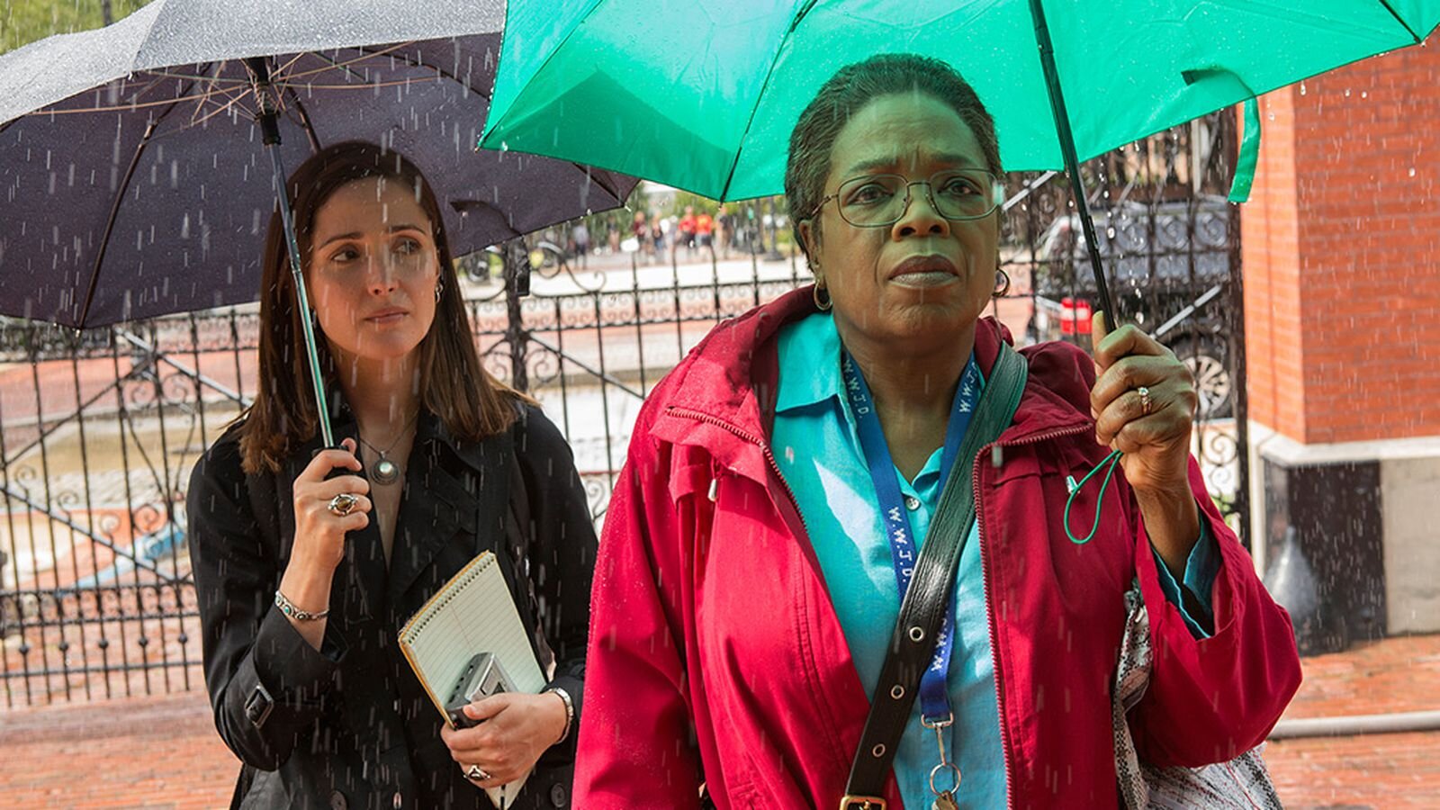 Two women standing outdoors in the rain, holding umbrellas, with a metal fence and brick building in the background. The woman in the foreground wears glasses, a red jacket, and has a serious expression. The woman in the background holds a purple umbrella and a notepad.