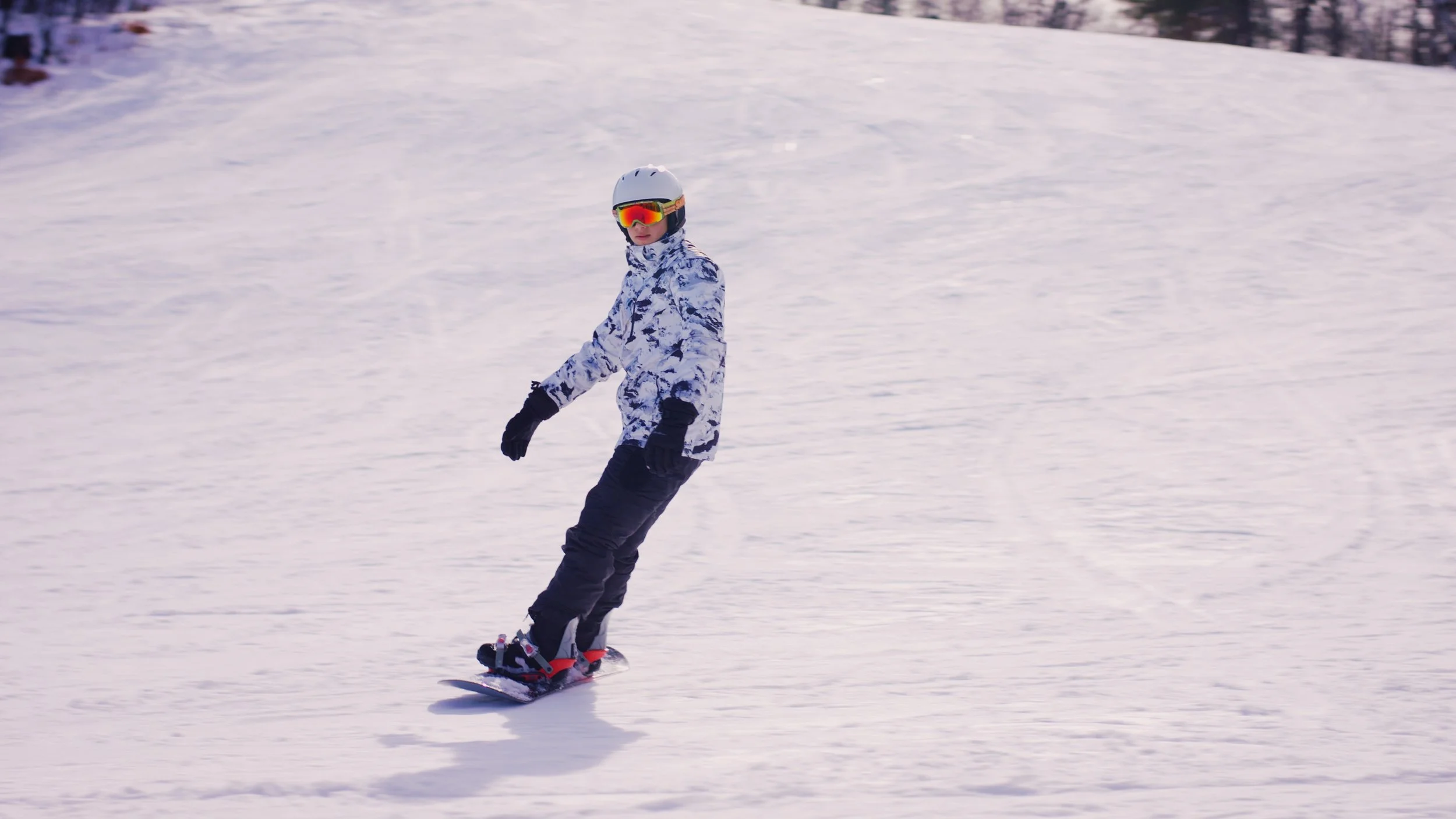 A person snowboarding on a snowy slope wearing a white helmet, reflective goggles, a white and blue patterned jacket, black gloves, and black snow pants.