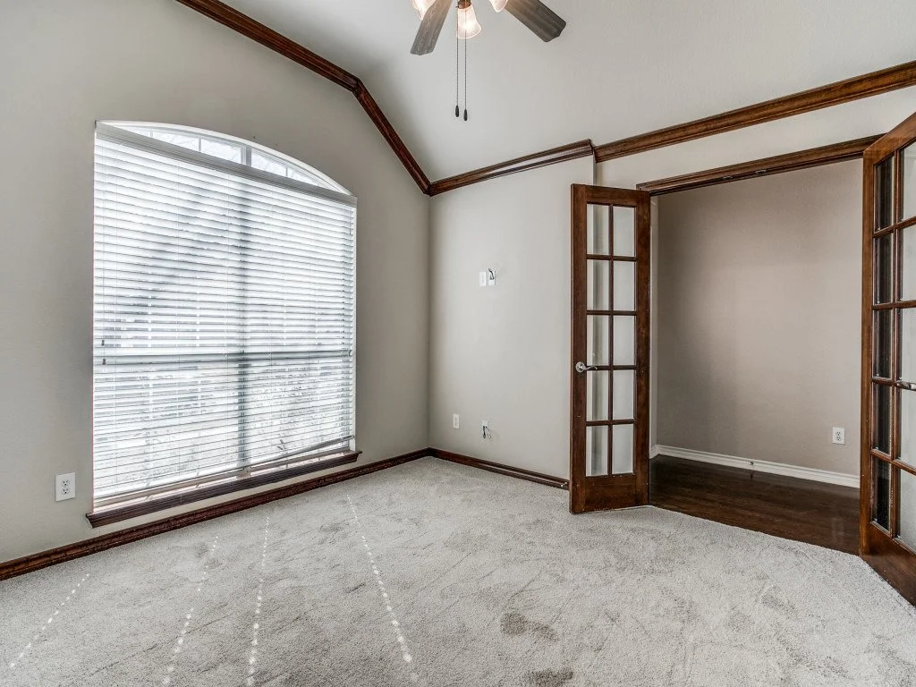 Empty room with large window with blinds, carpeted floor, wood trim, and French doors leading to another space.