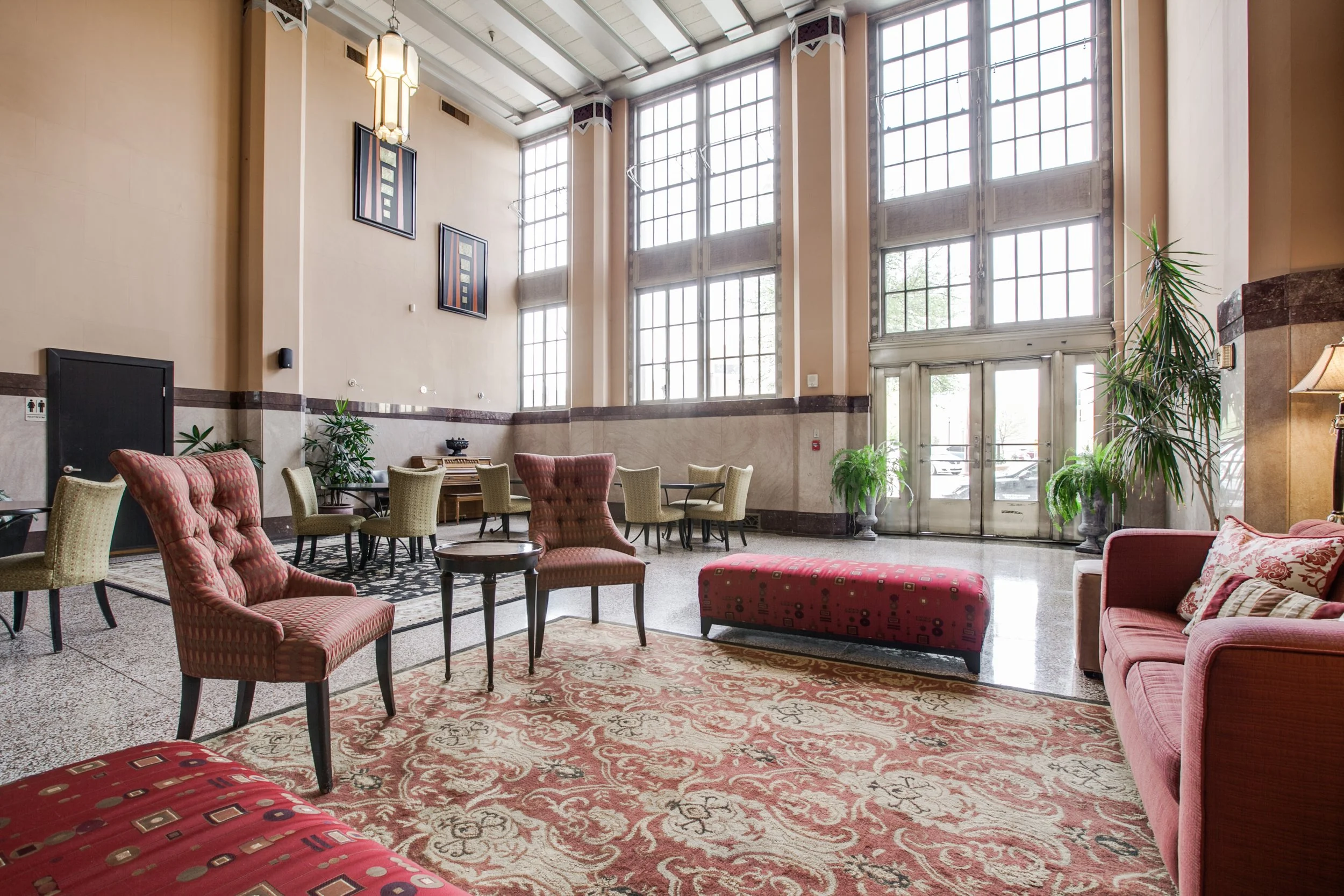 A spacious hotel lobby with large windows, vintage furniture including pink and beige armchairs, a pink upholstered bench, green potted plants, and wall artwork.