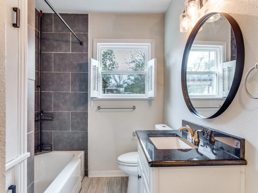 Bathroom with a bathtub, shower, toilet, vanity with black countertop, round mirror, window with shutters, and light fixture.
