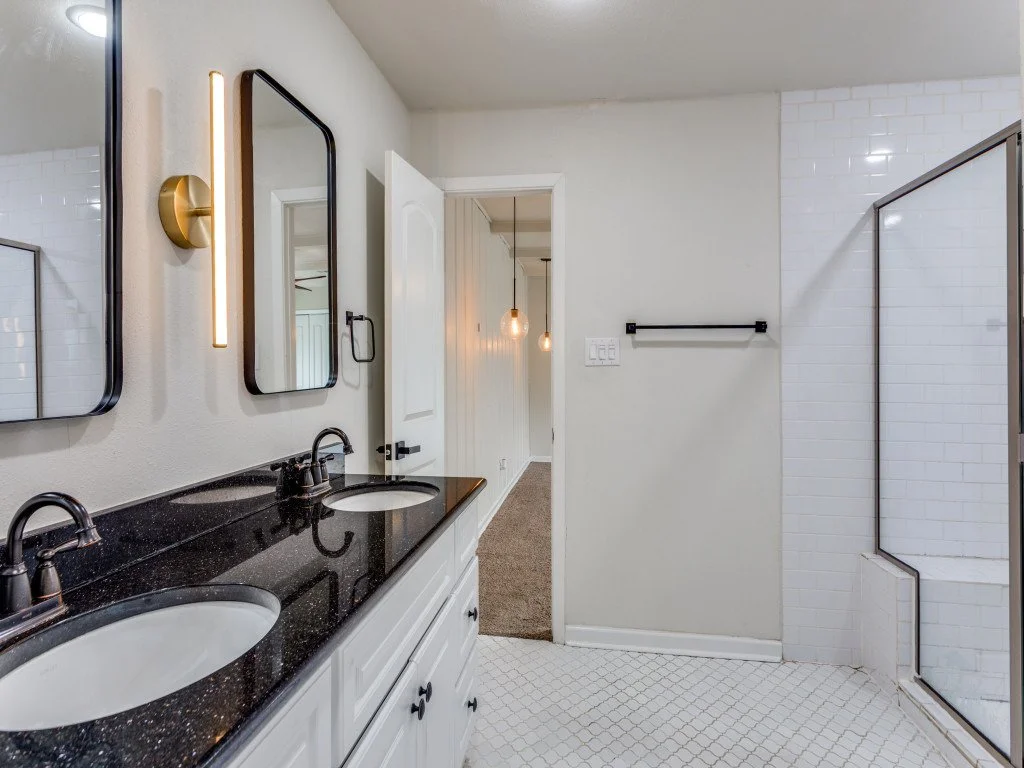 Bathroom with a black vanity countertop, two sinks, black faucets, three mirrors, a gold wall light, a white tiled shower with a glass door, white walls, a towel rack, and a doorway leading to another room with hanging pendant lights.