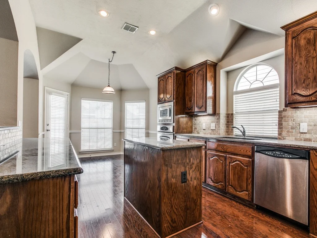 Kitchen with wooden cabinets, granite countertops, a dishwasher, a microwave, a window with blinds, and a small island in the center.