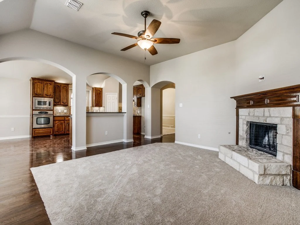 Empty living room with a fireplace, ceiling fan, and open kitchen area with wooden cabinets and stainless steel appliances.