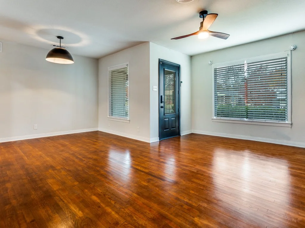Empty living room with hardwood floors, white walls, two large windows with blinds, a black front door, a ceiling fan, and a modern hanging light fixture.