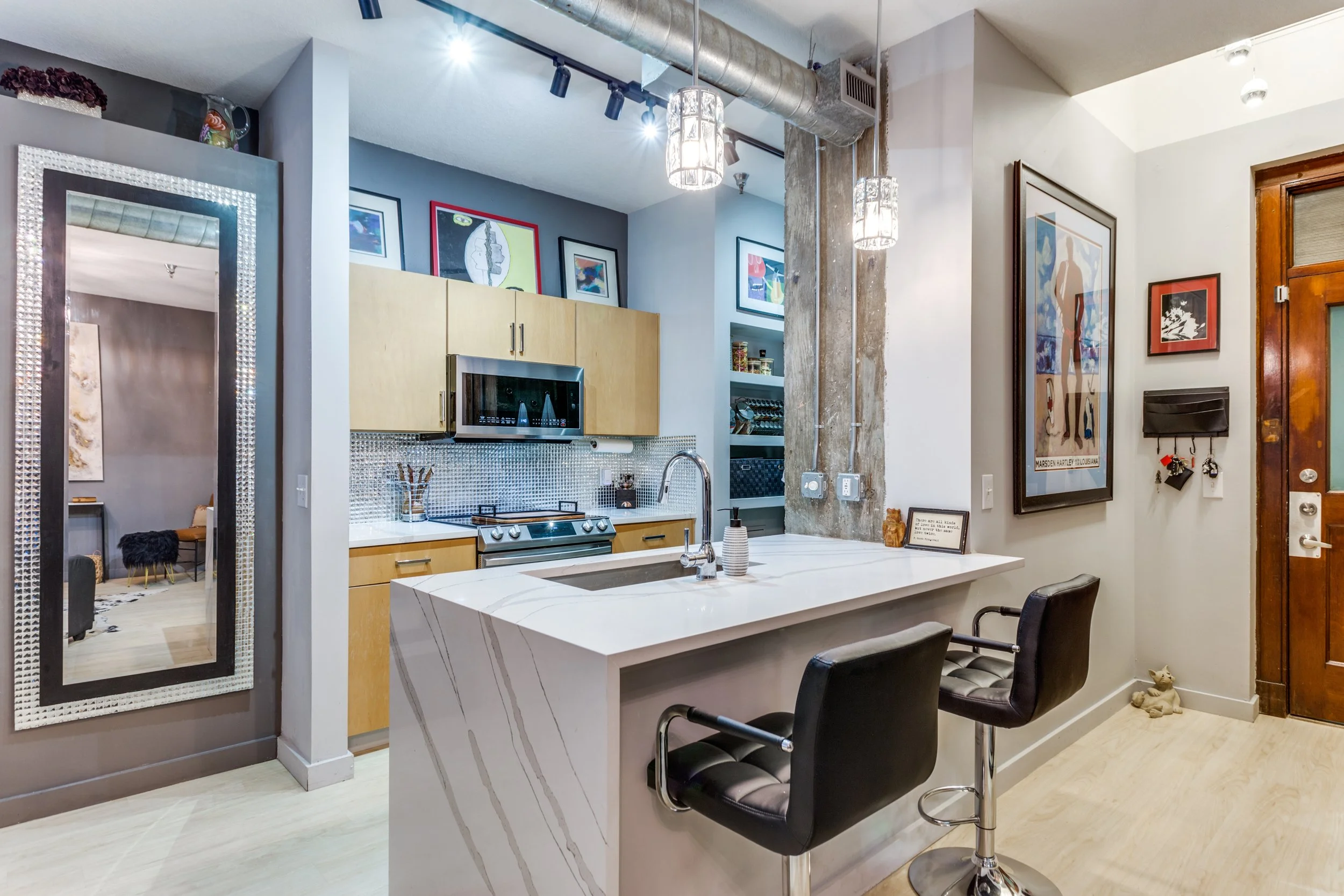 Modern kitchen with white marble island, black bar stools, light wood cabinets, stainless steel appliances, artwork on the walls, and a large mirror.